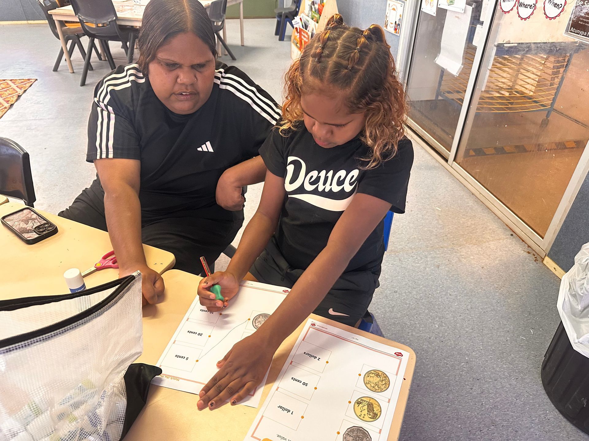 Two children lean over a table, writing on papers with markers in a bright indoor space.