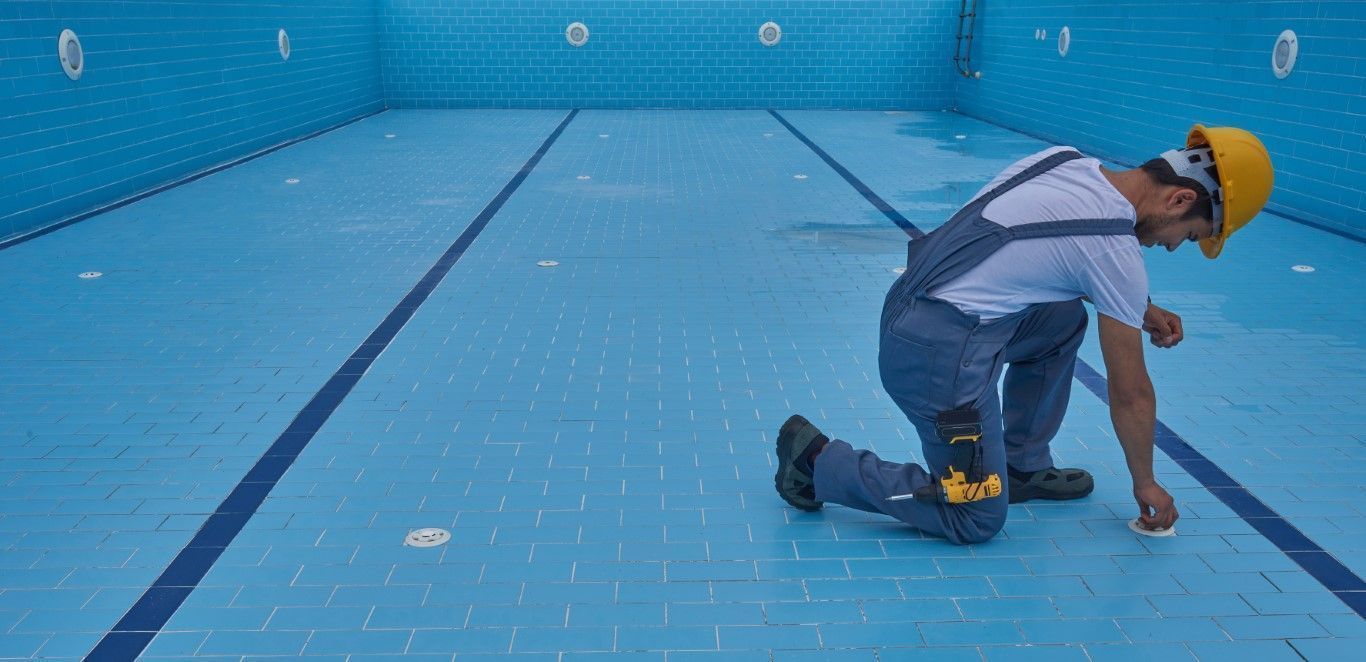Worker in yellow hard hat kneeling in an empty blue swimming pool, inspecting the floor.