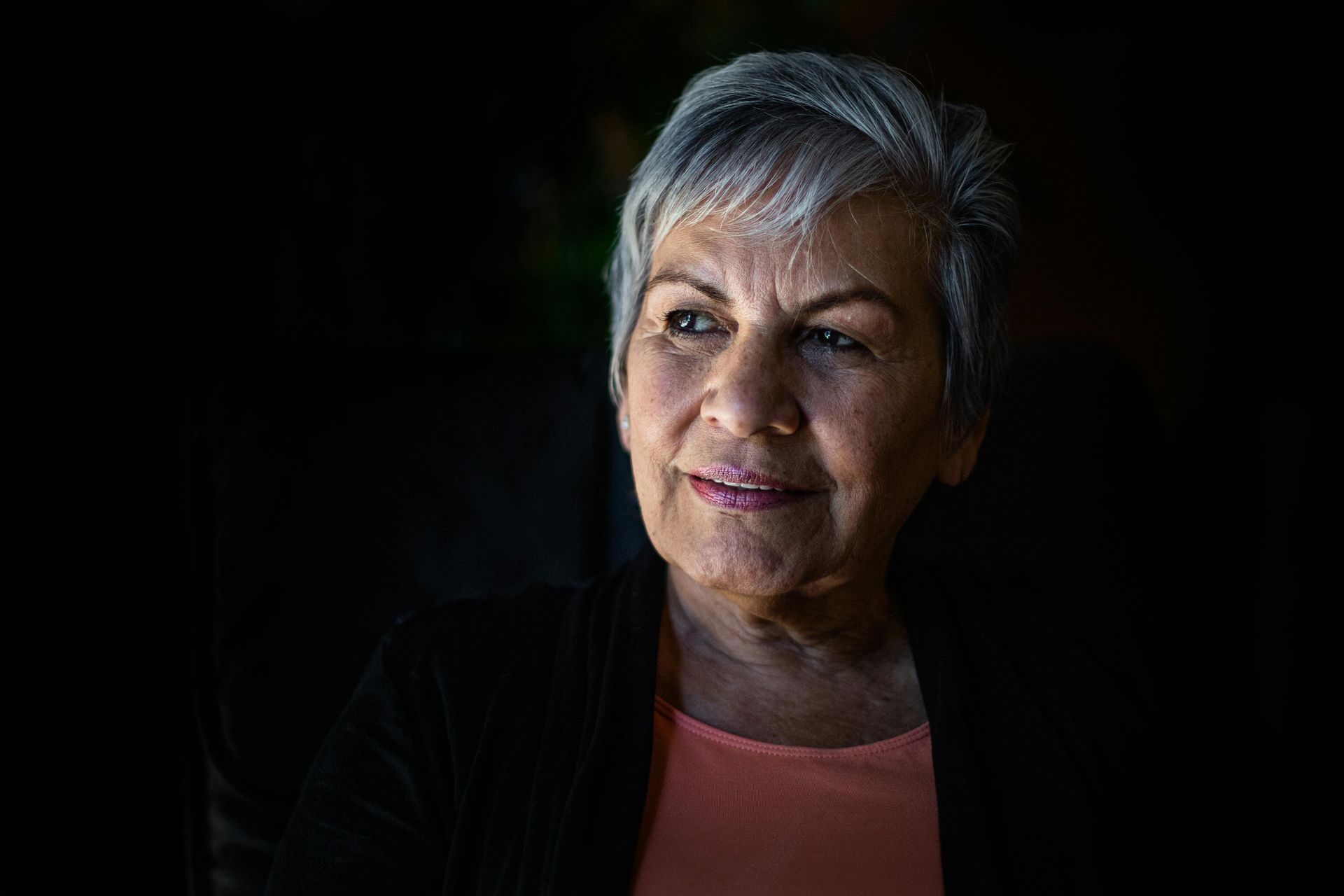 An elderly woman with gray hair is smiling in a dark room.
