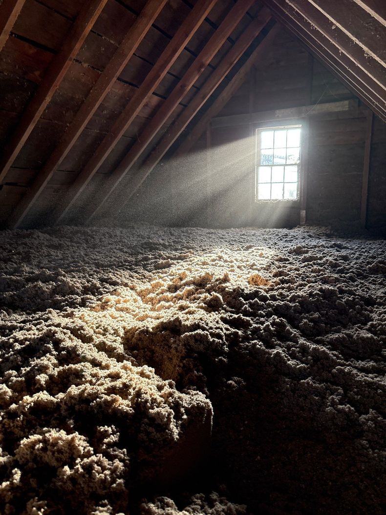 Attic with slanted wooden beams, sunbeams through a window, and piles of insulation on the floor