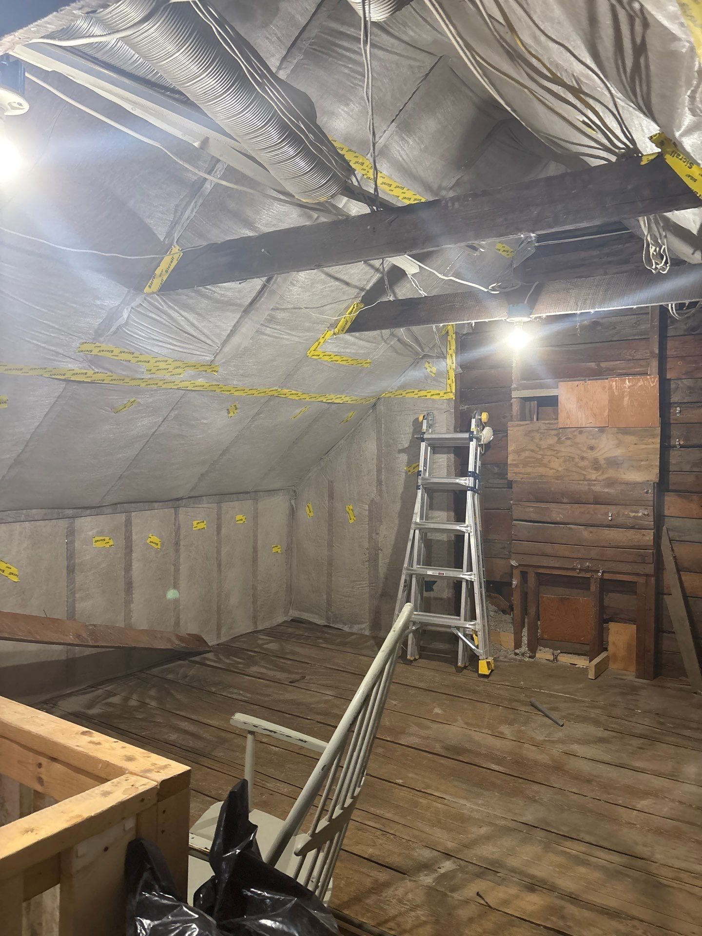 Attic under renovation with exposed insulation, ladder, wooden floor, and construction materials.