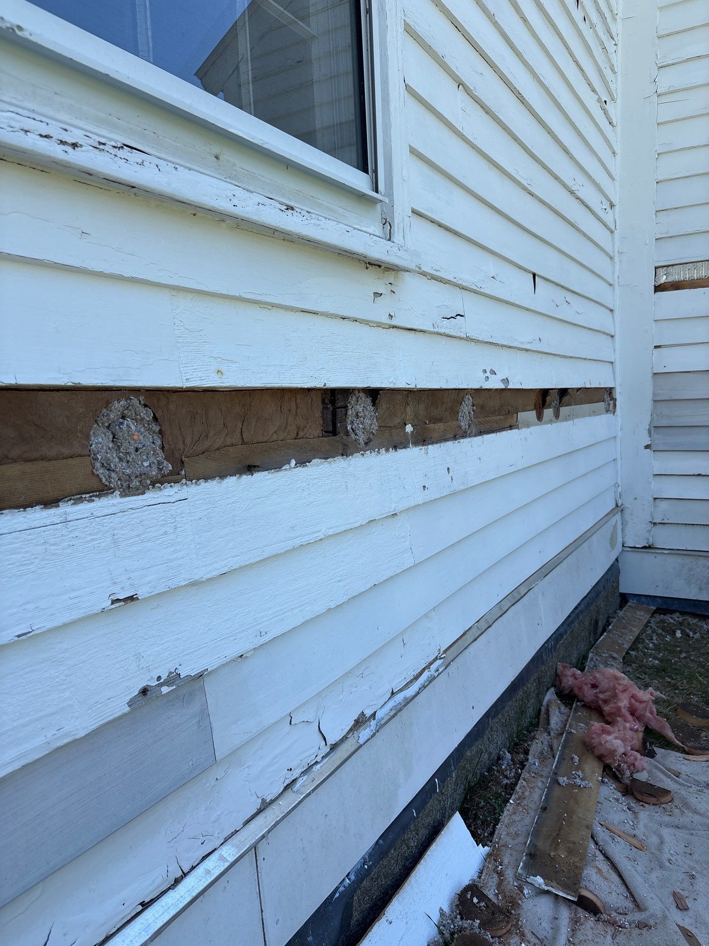 Damaged white house siding with exposed wood and debris along the foundation