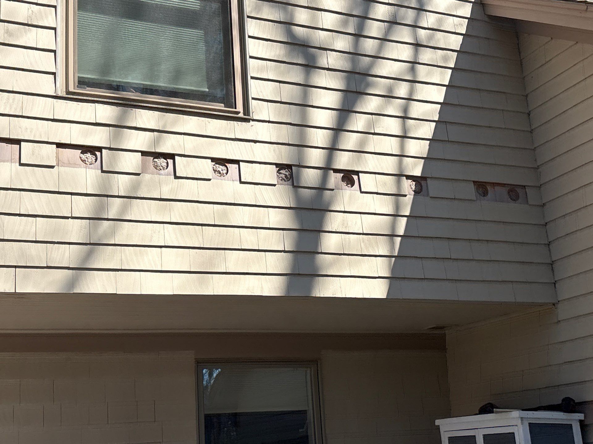 Beige house exterior with a second-story window and a row of exposed wall patches under the eaves.