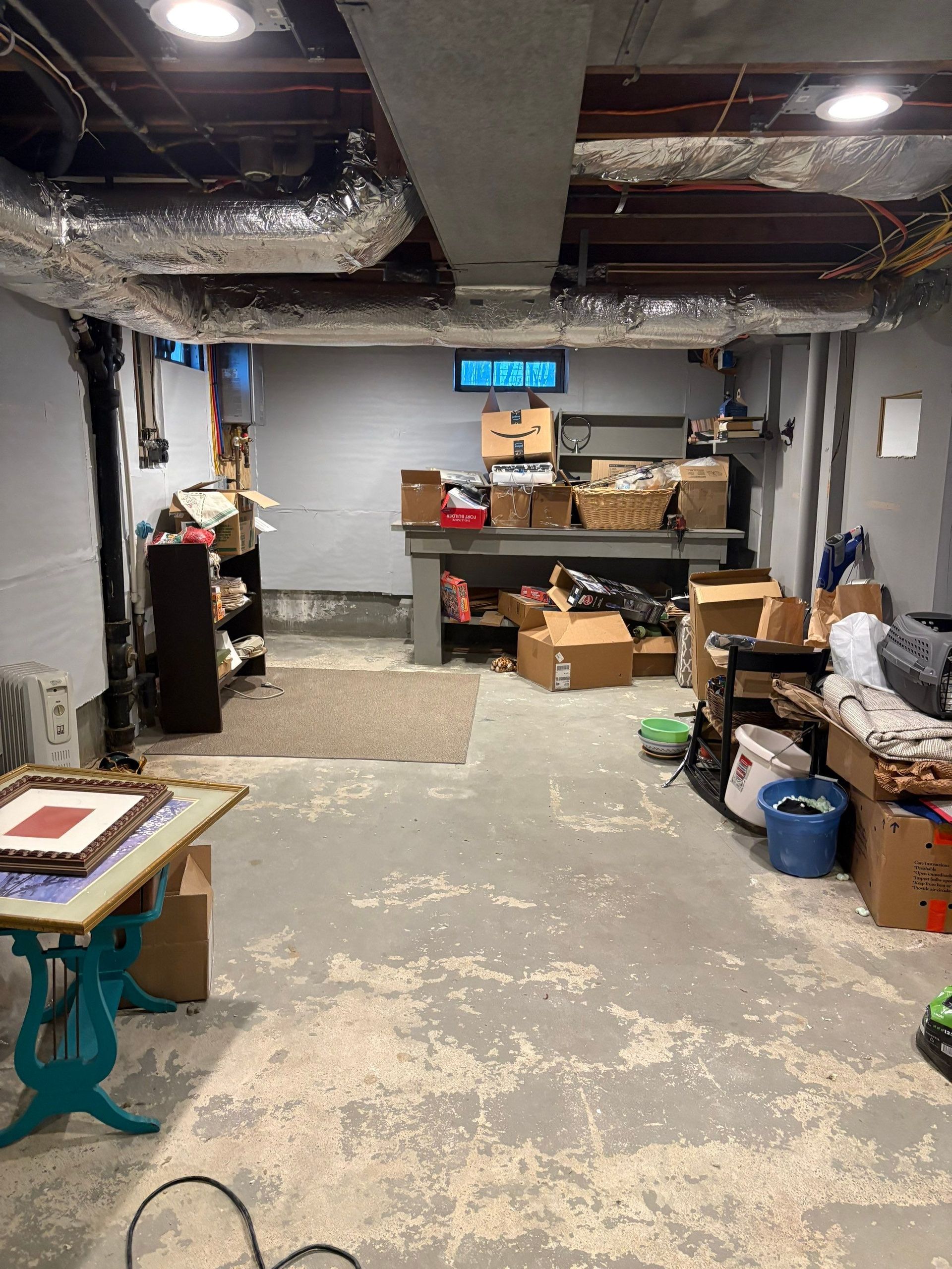 Cluttered basement workspace with tools, boxes, shelves, and a concrete floor under exposed ductwork.