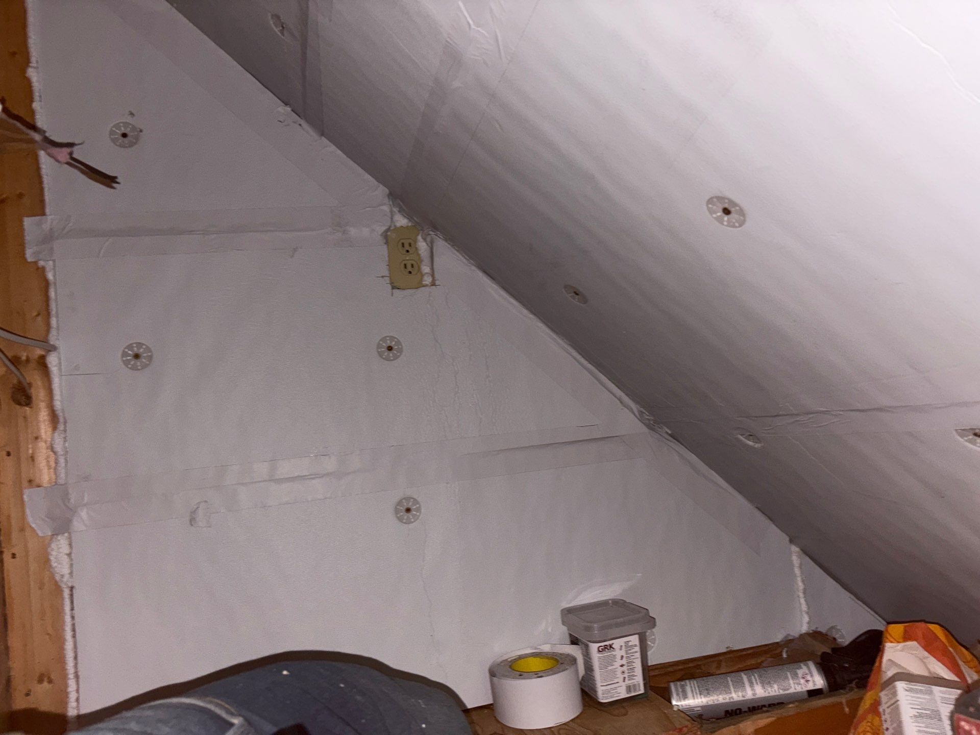 Attic corner with slanted white walls, exposed wood beam, and a few paint cans and tools on the floor