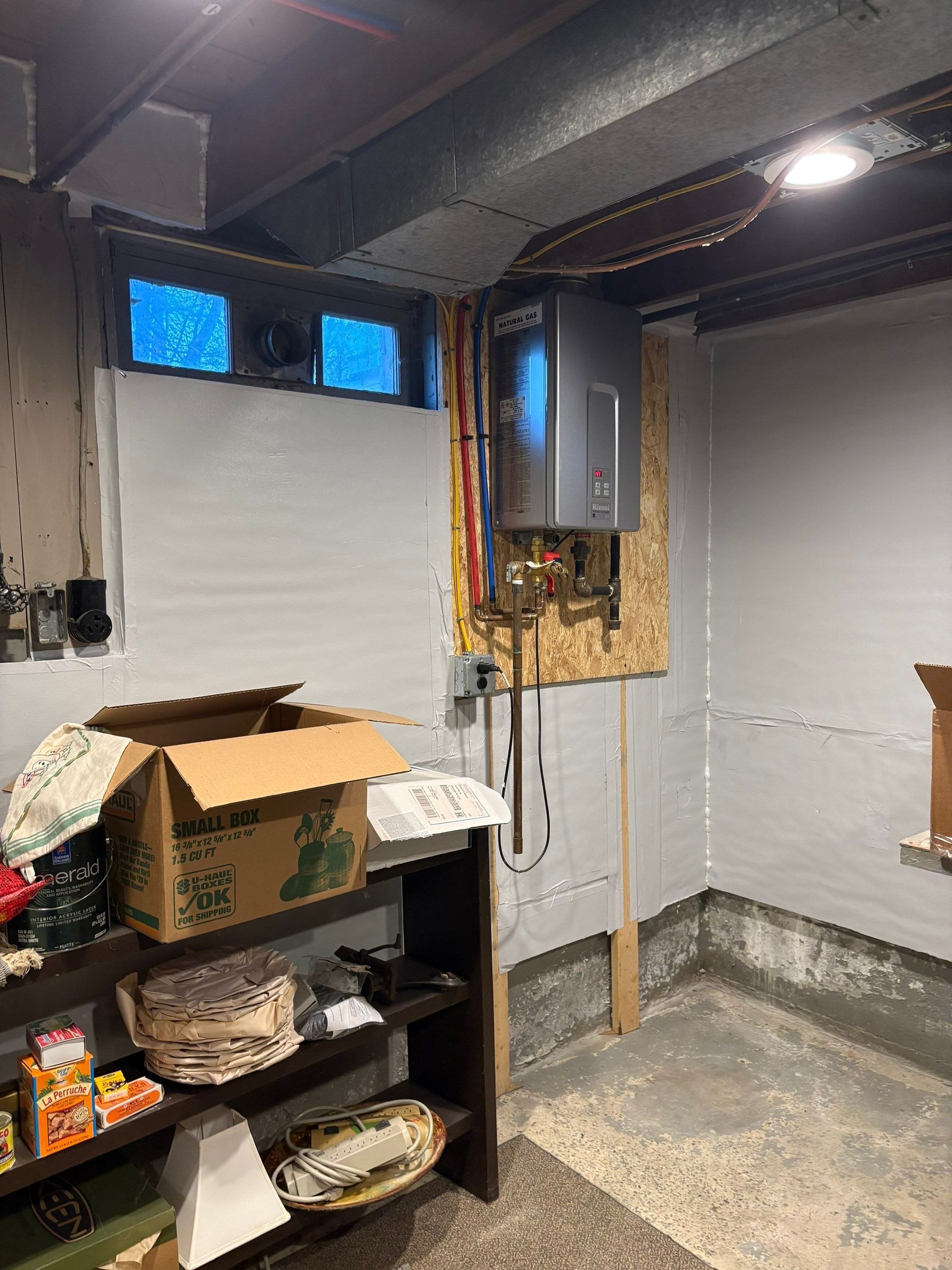 Basement utility room with shelves, cardboard boxes, and exposed pipes near a small window.