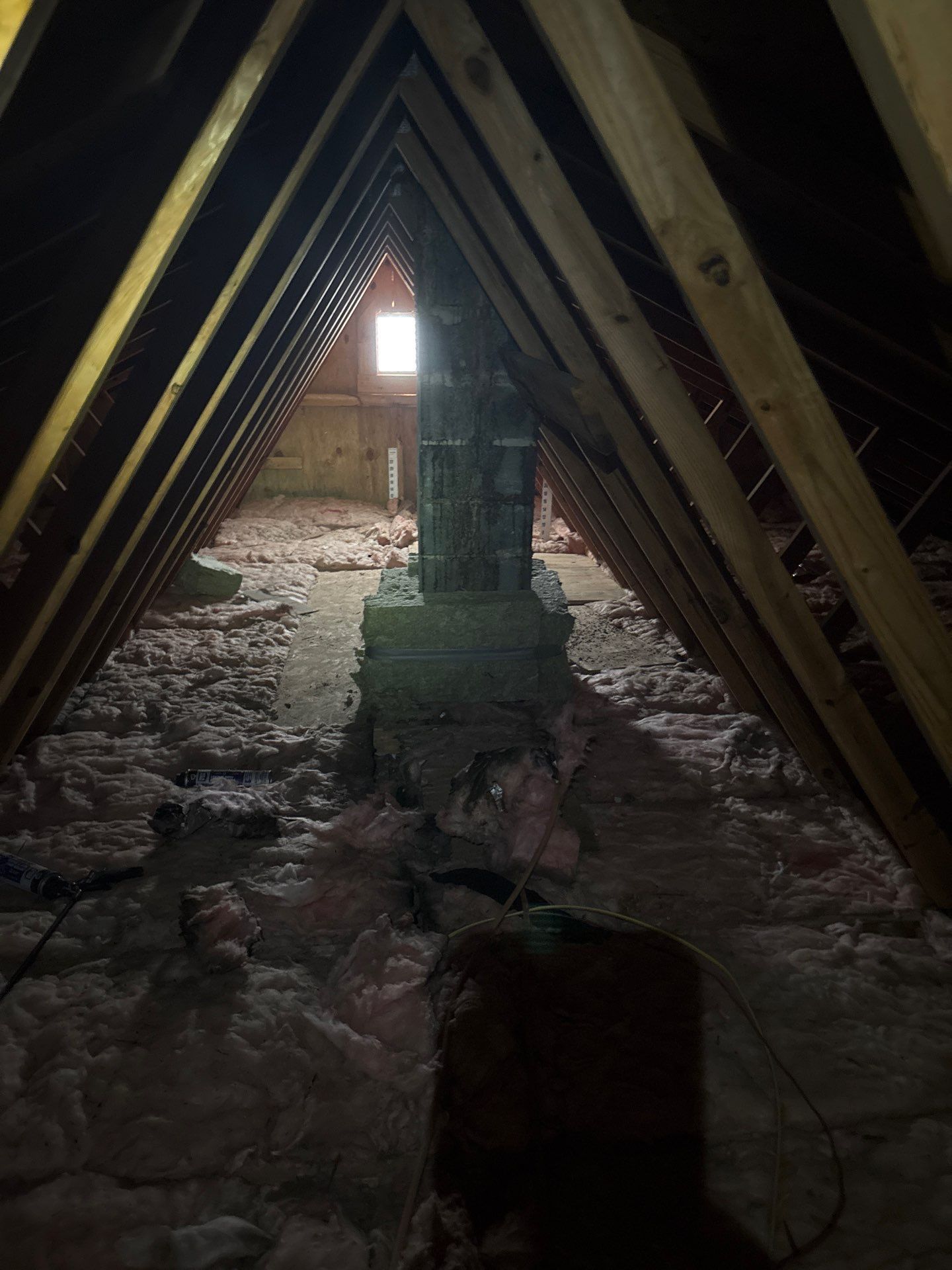 Attic with exposed rafters, insulation, and a central chimney or vent shaft, lit by a small rear window.