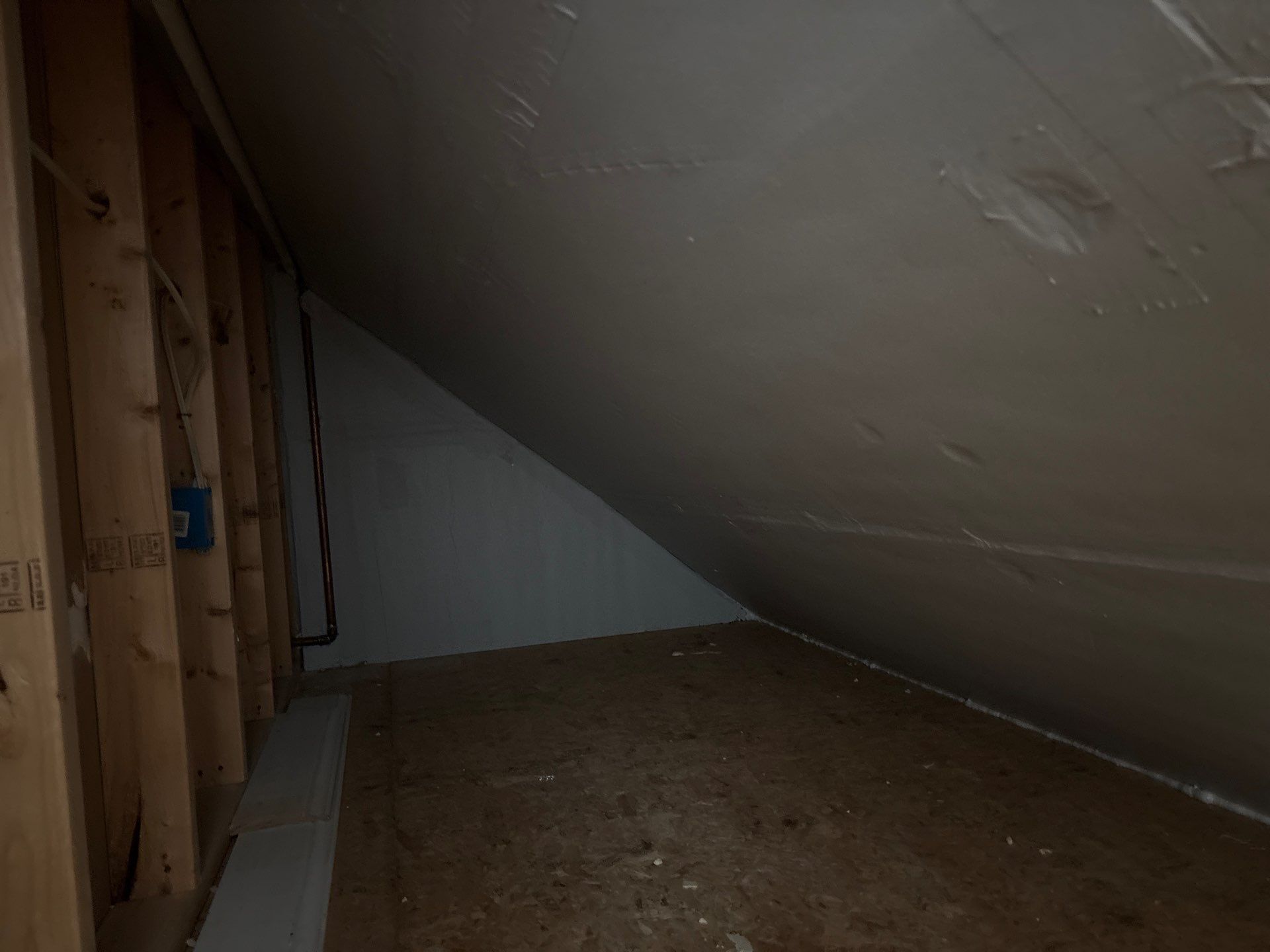 Small unfinished attic room with slanted white ceiling, exposed wooden framing, and bare plywood floor.