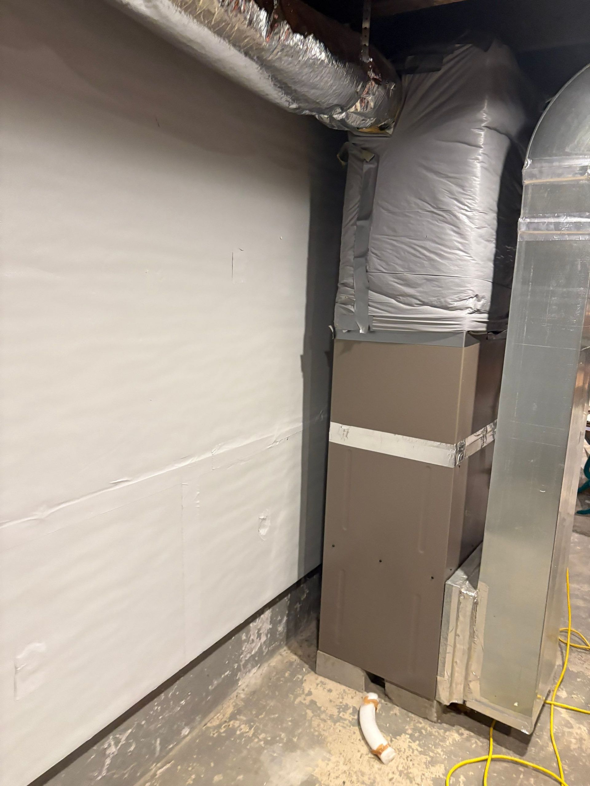 Basement corner with insulated ductwork, white wall, and exposed piping on a concrete floor
