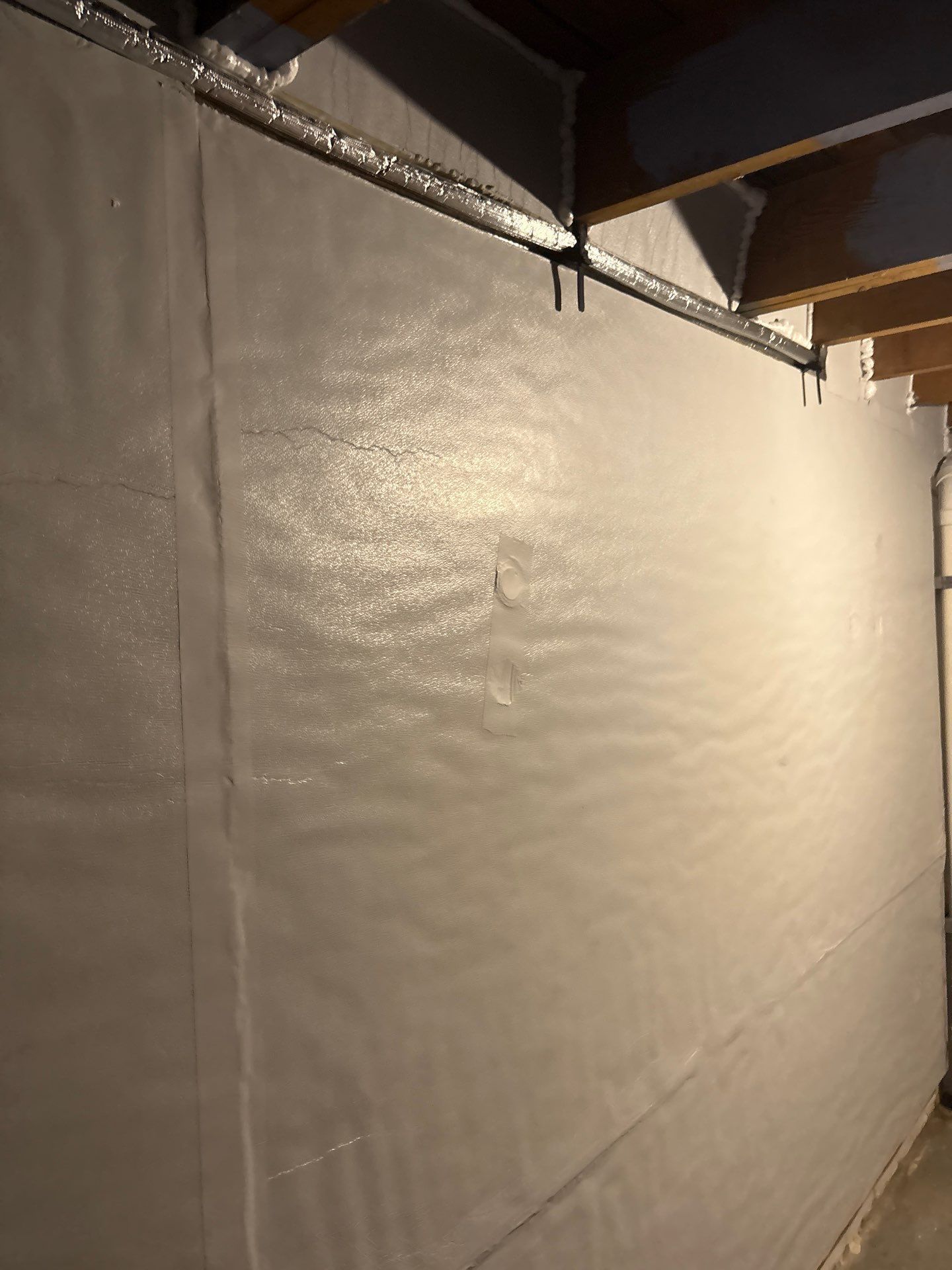 Dimly lit unfinished wall with beige insulation or tarp beneath exposed ductwork and ceiling beams