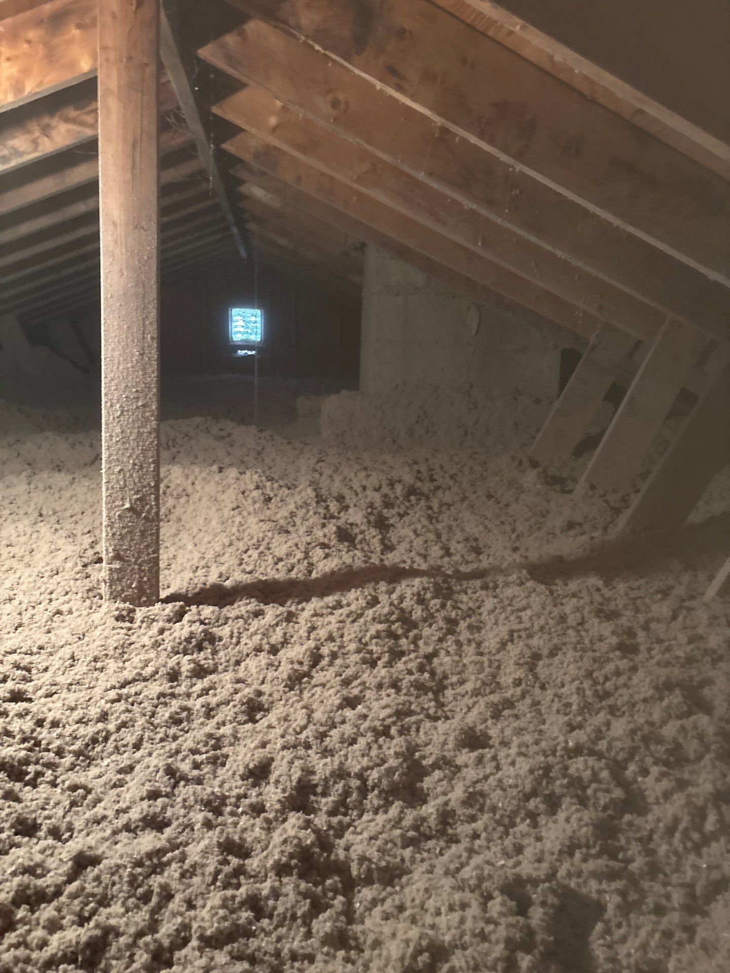 Attic space with exposed beams and insulation-covered floor, lit by a small window in the distance.
