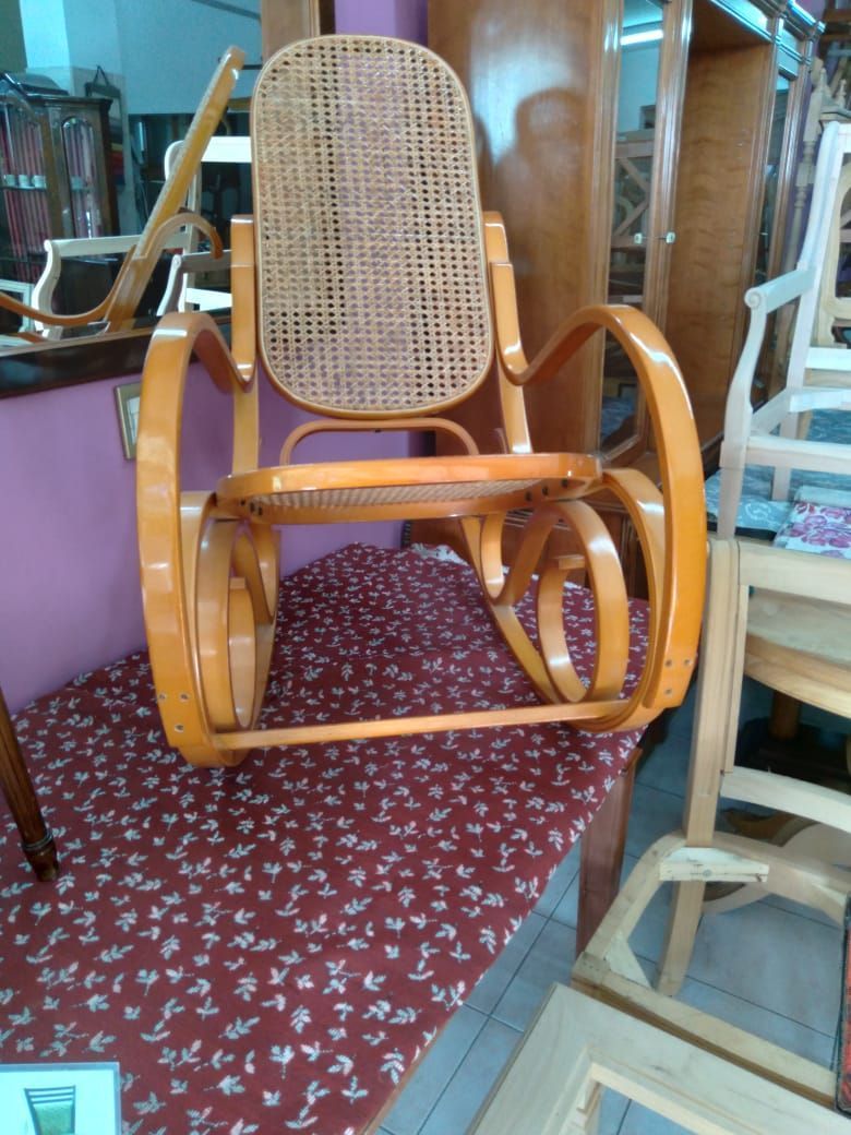 Mecedora de madera con respaldo y asiento tejidos sobre una alfombra roja estampada en una tienda de muebles.
