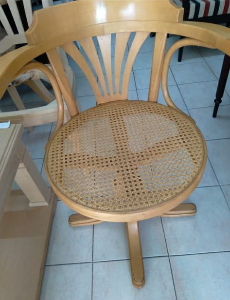 Silla de madera clara con asiento tejido y respaldo redondeado, fotografiada en interiores sobre un suelo de baldosas.