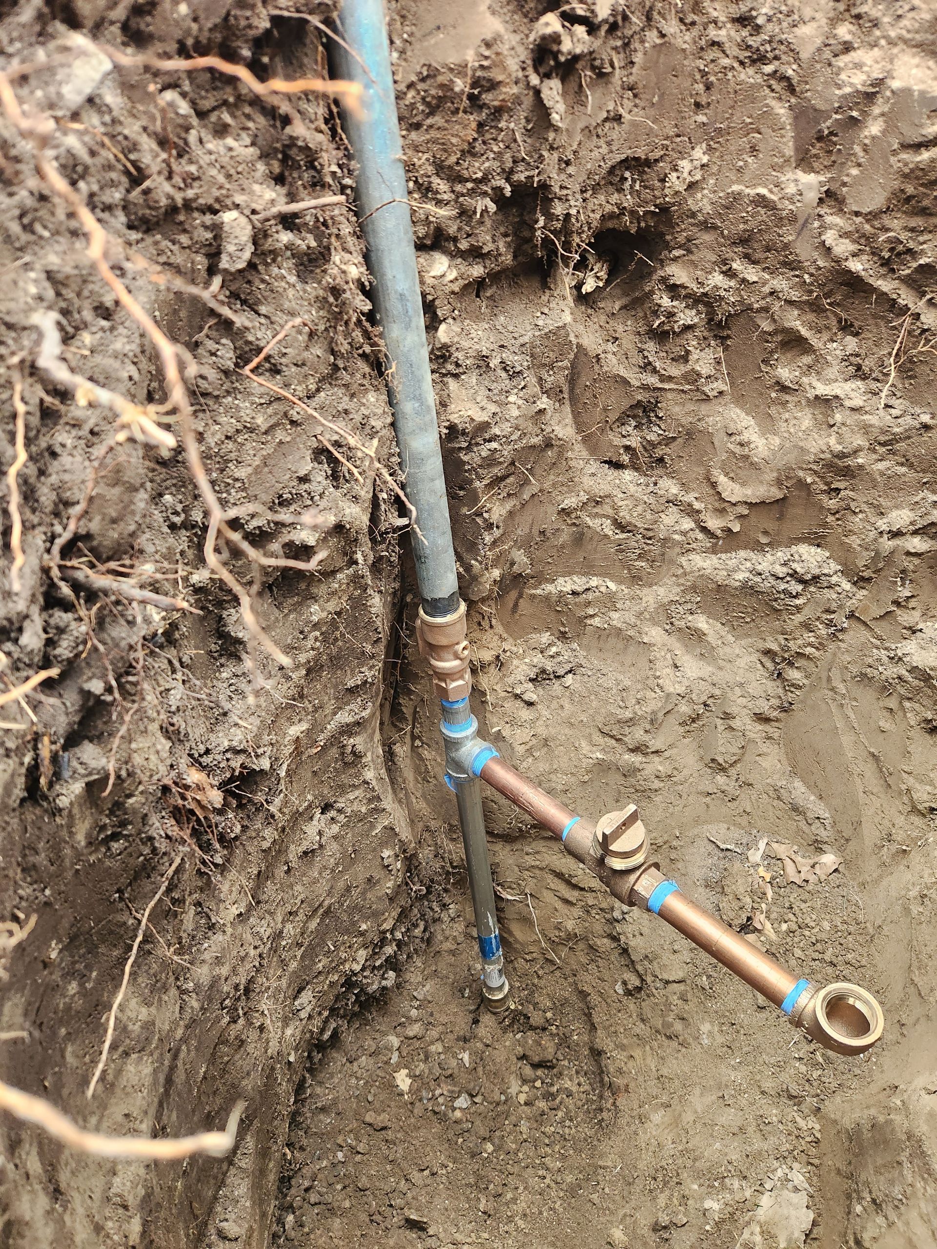 A blue pipe and copper pipes with fittings and a spigot, installed in a trench dug in dirt.