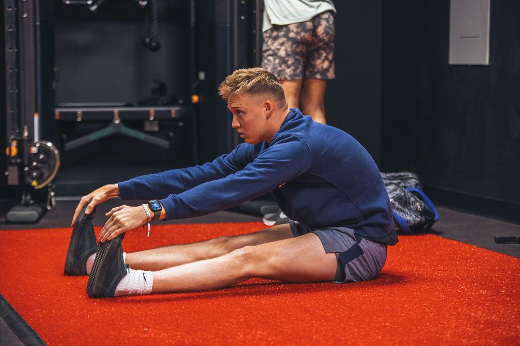 A man is sitting on the floor stretching his legs in a gym.