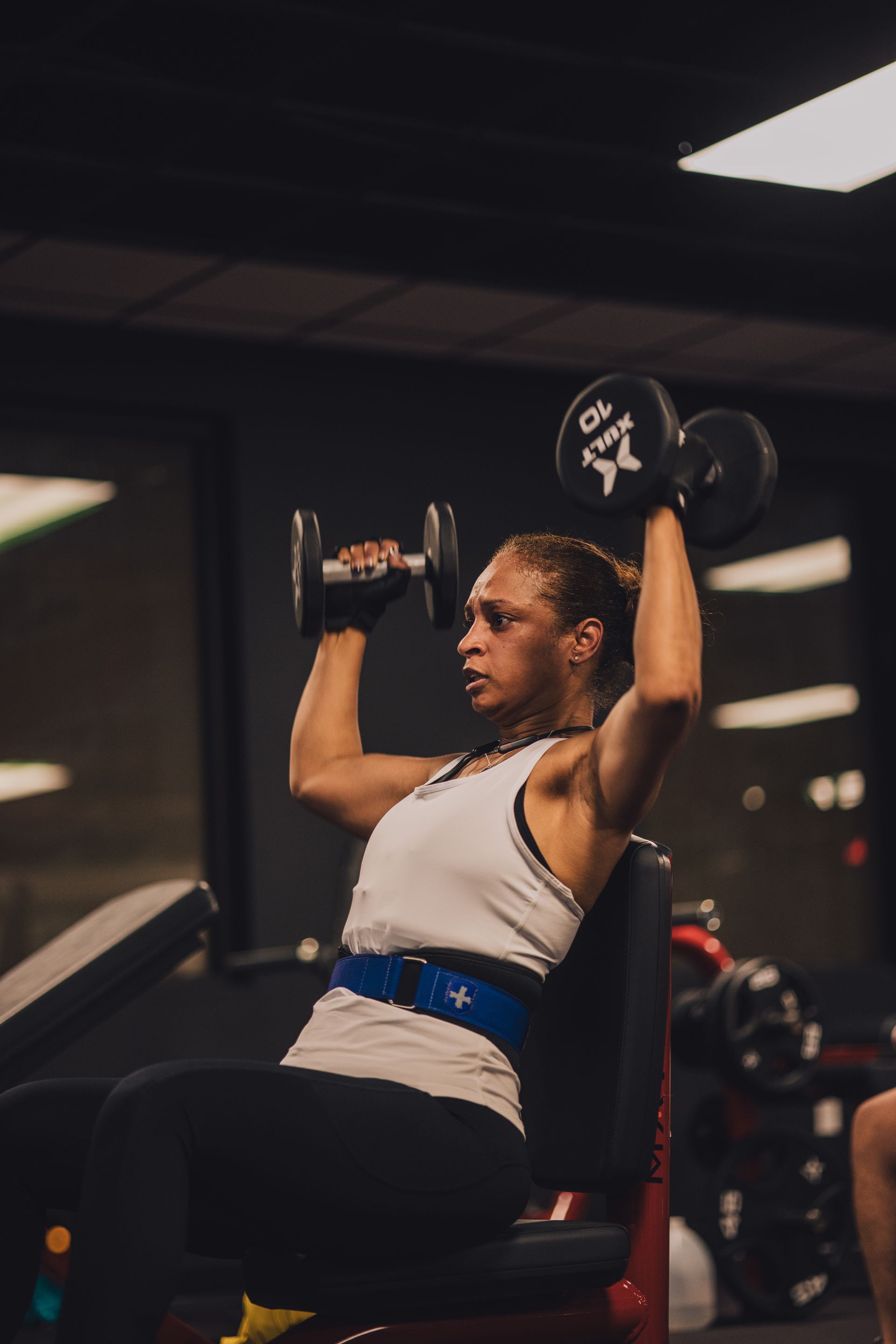 A woman is lifting a dumbbell while sitting on a bench in a gym.