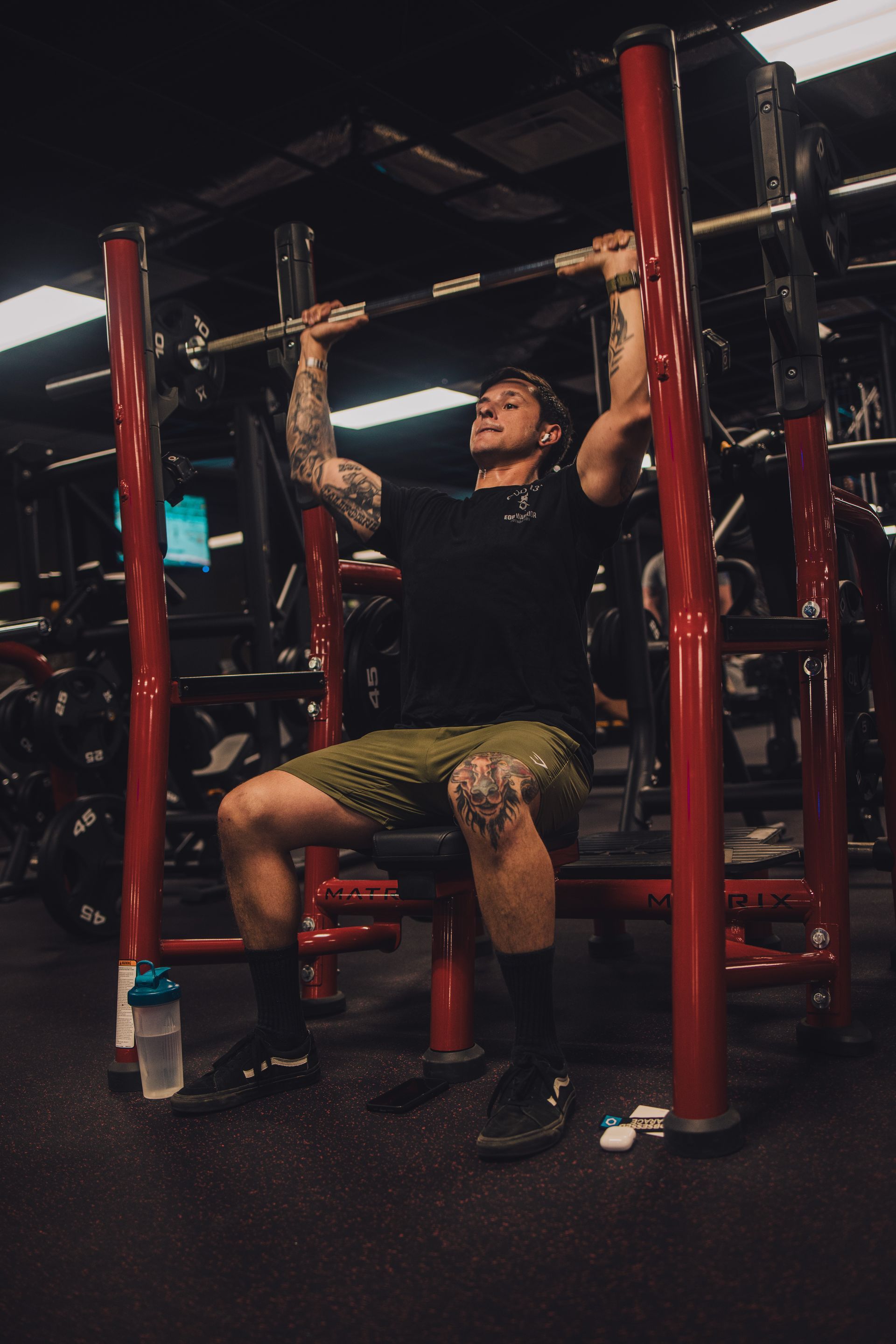 A man is sitting on a bench in a gym holding a barbell over his head.