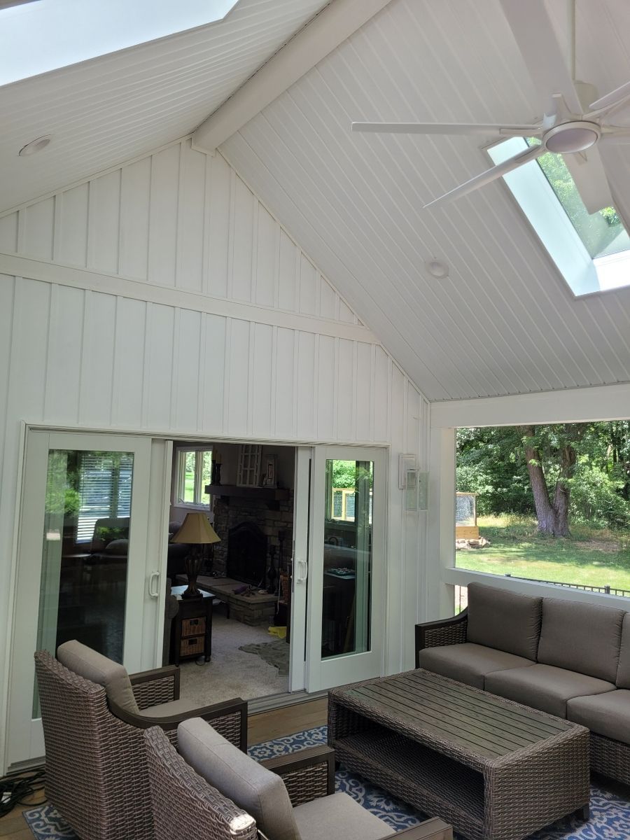 Sunroom with white paneling, skylight, and open doors to a living room, furnished with wicker seating and a coffee table.