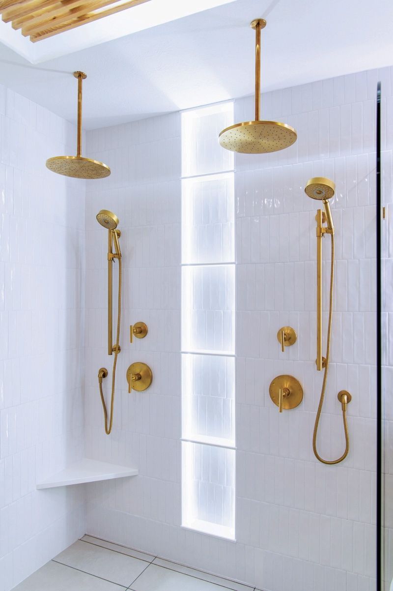 Two gold showerheads and fixtures on white tiled walls. Vertical recessed lit shelf.