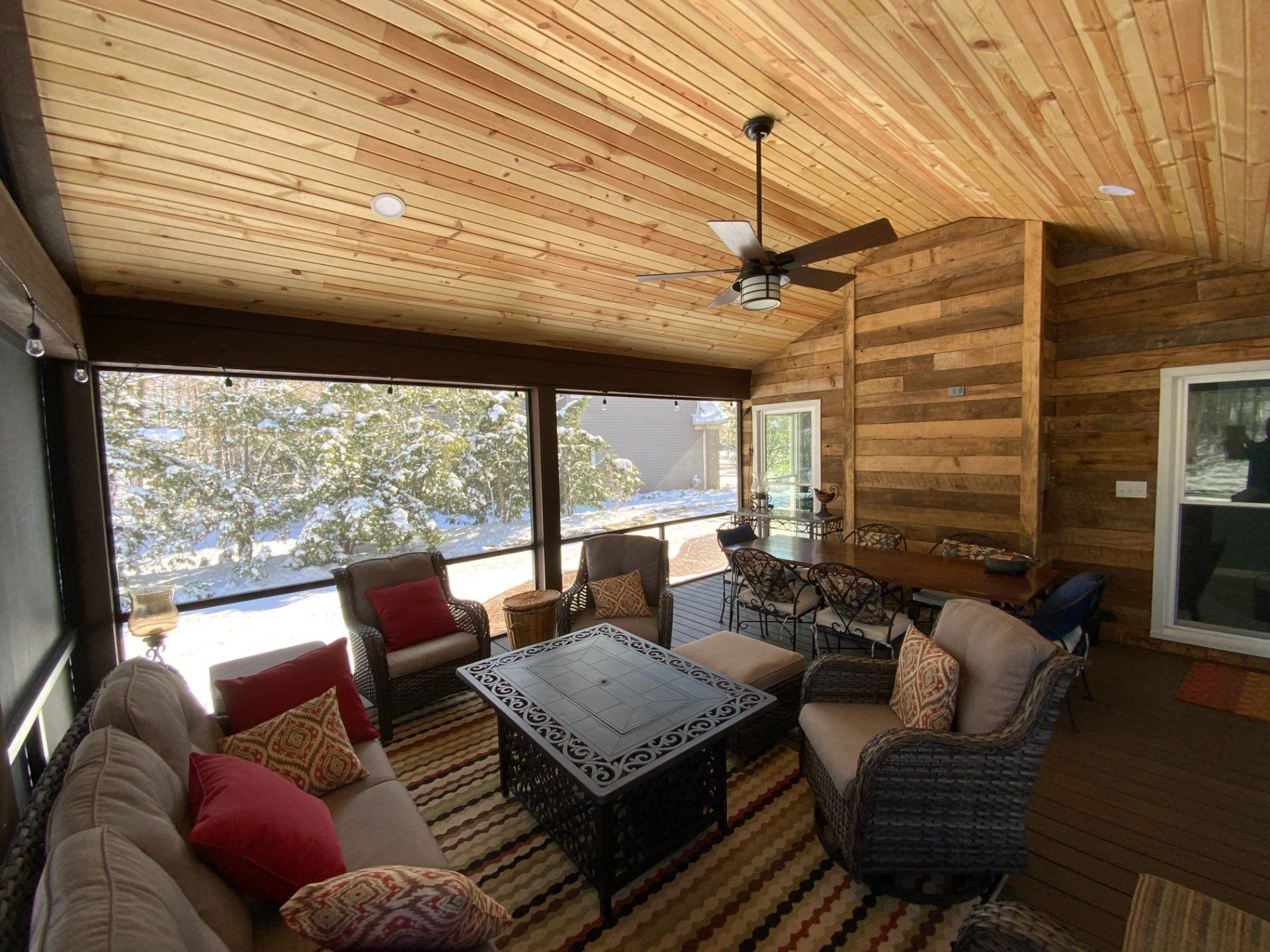 Enclosed patio with wood ceiling, walls, and screens. Furnished with seating, rug, and fire pit, overlooking snowy trees.