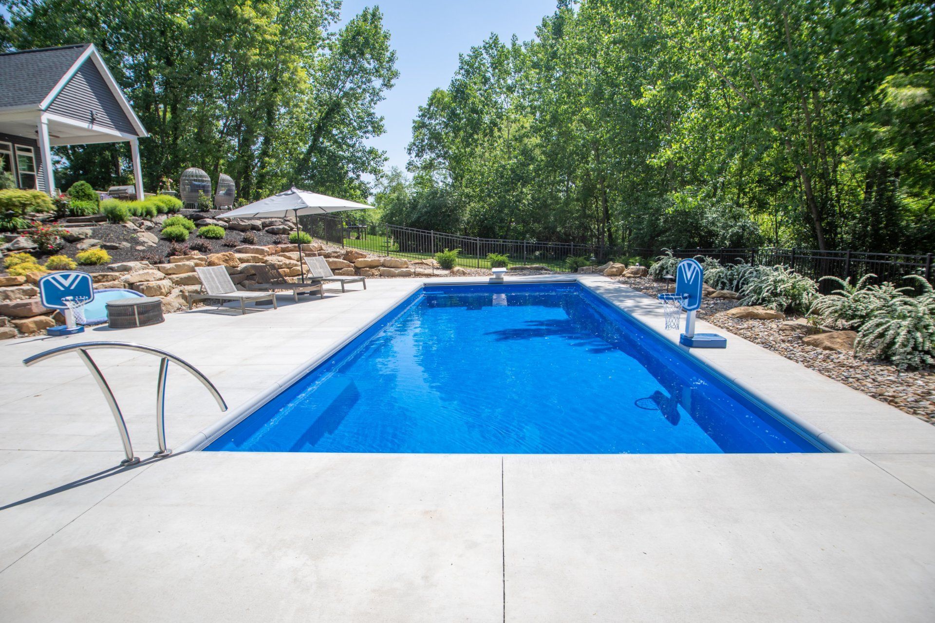 Rectangular in-ground pool surrounded by concrete patio, with trees and a house in the background. Blue water.