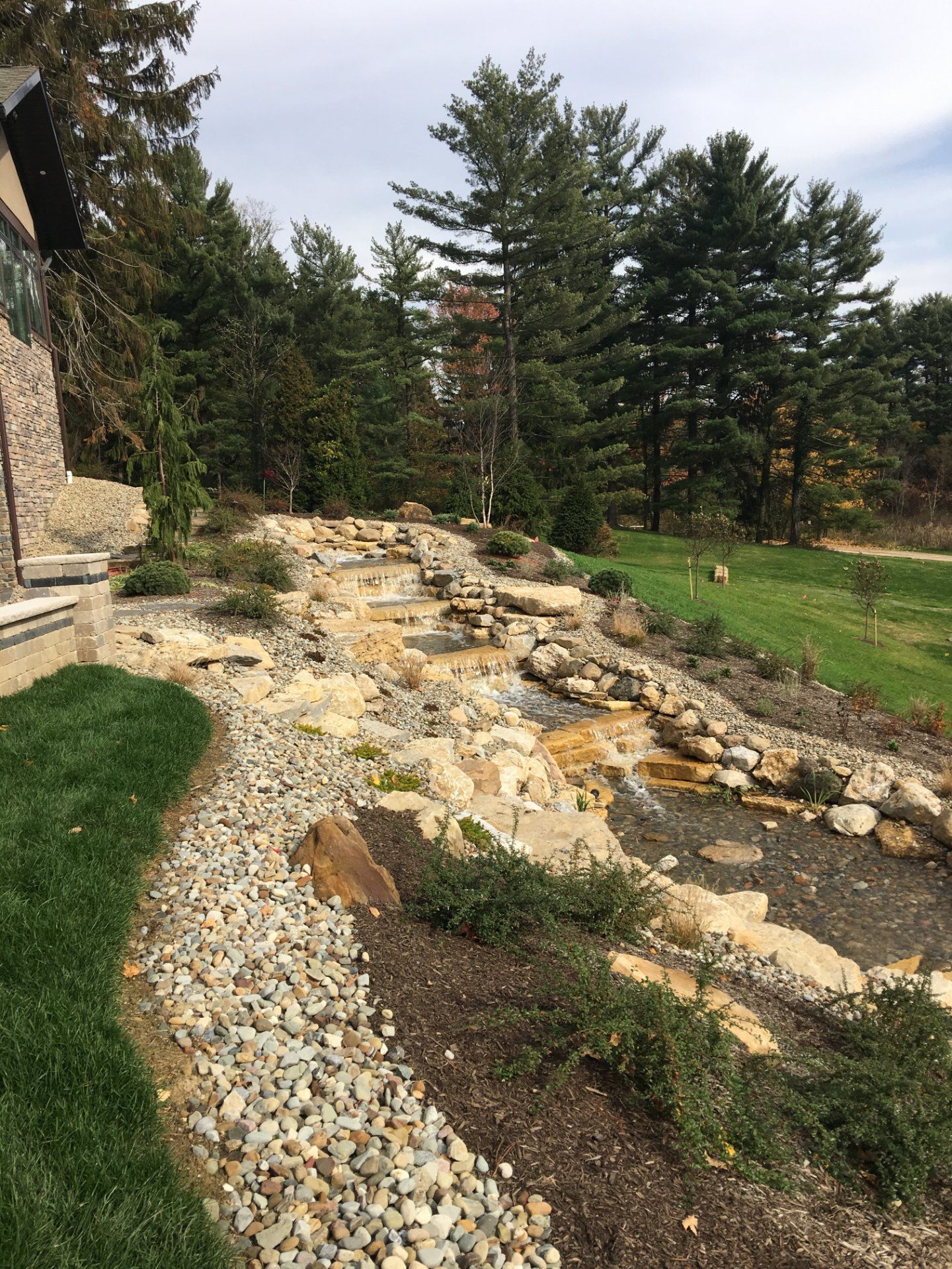 Stone waterfall feature with greenery and landscaping beside a home with tall trees in the background.
