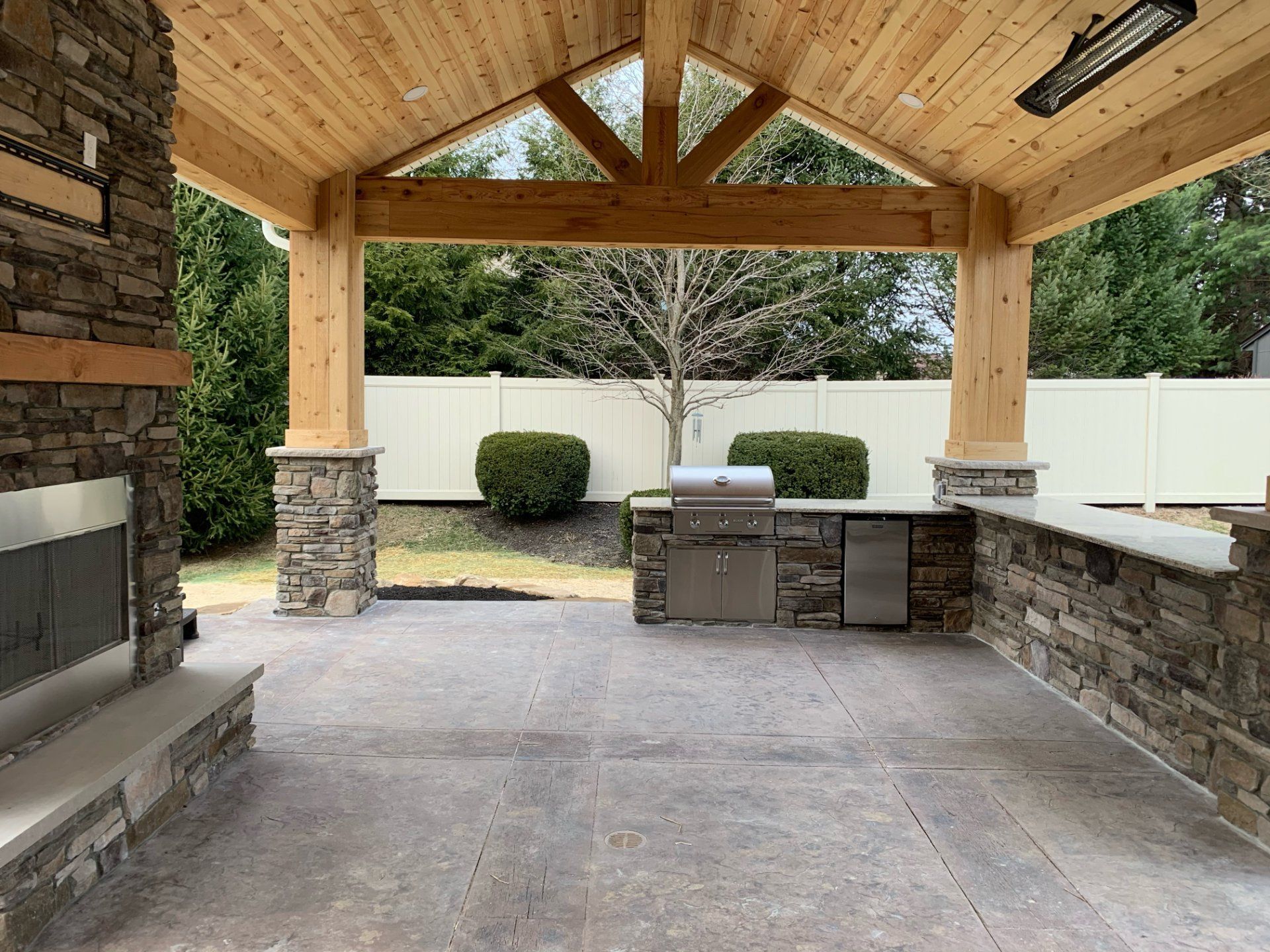 Outdoor kitchen with stone fireplace and countertop, wood beam ceiling, stainless steel grill, and white fence.