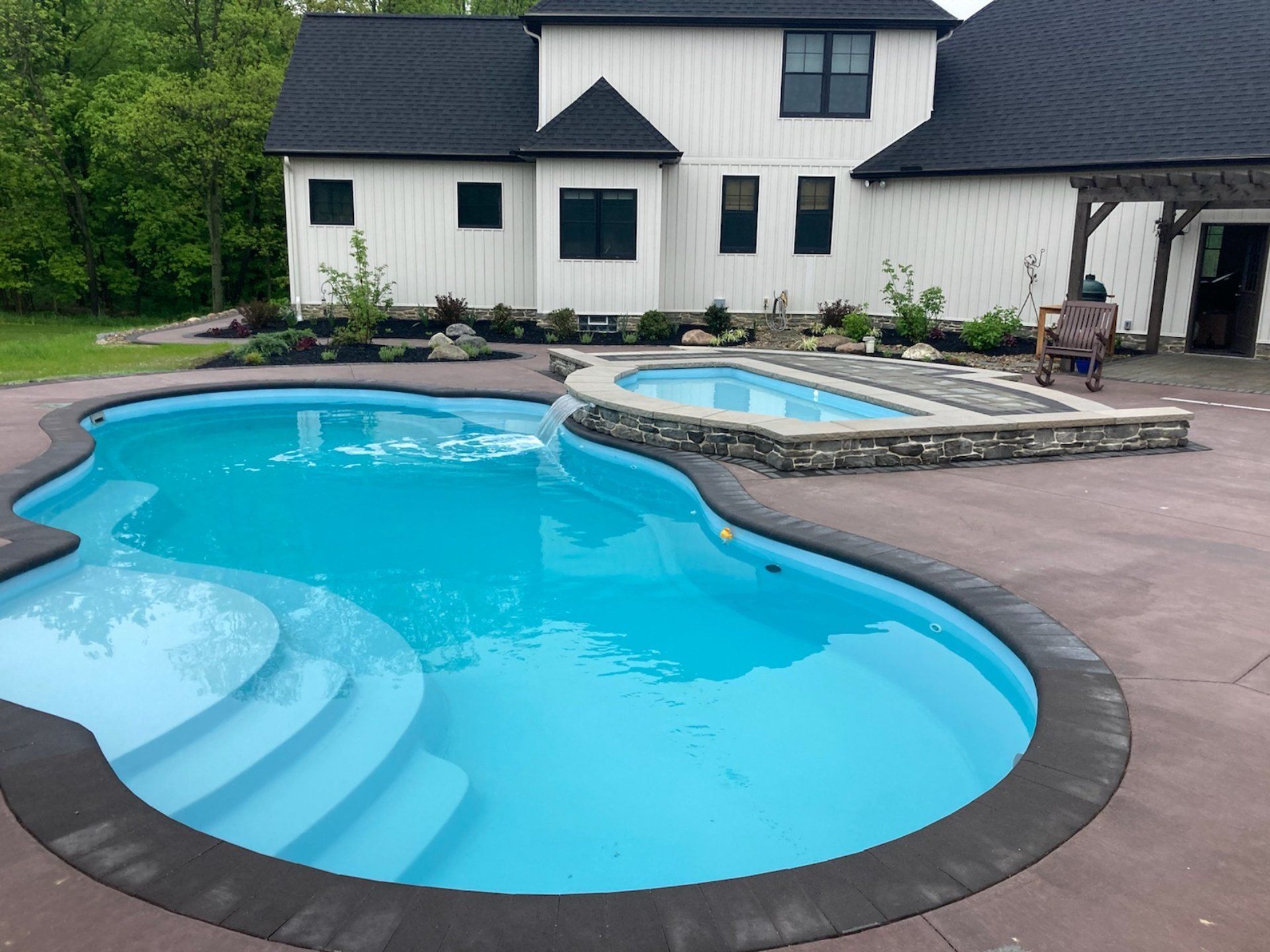 A backyard with a blue-water pool, a hot tub, and a white house in the background. The pool has steps and a dark border.