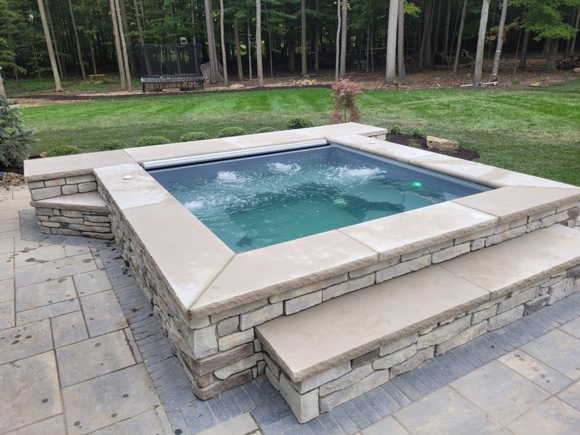 Stone-bordered, square spa with bubbling water, on a patio with steps. Green lawn and trees in the background.