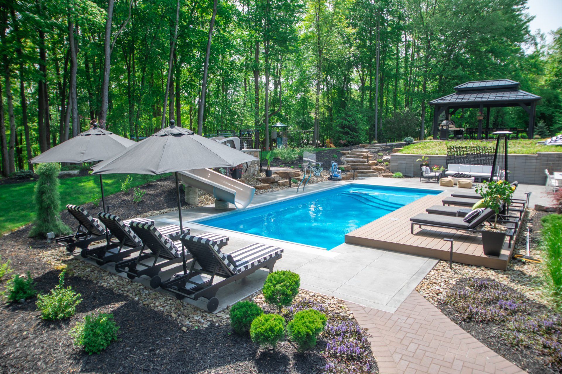 Pool area with lounge chairs, umbrellas, and a gazebo, surrounded by trees.