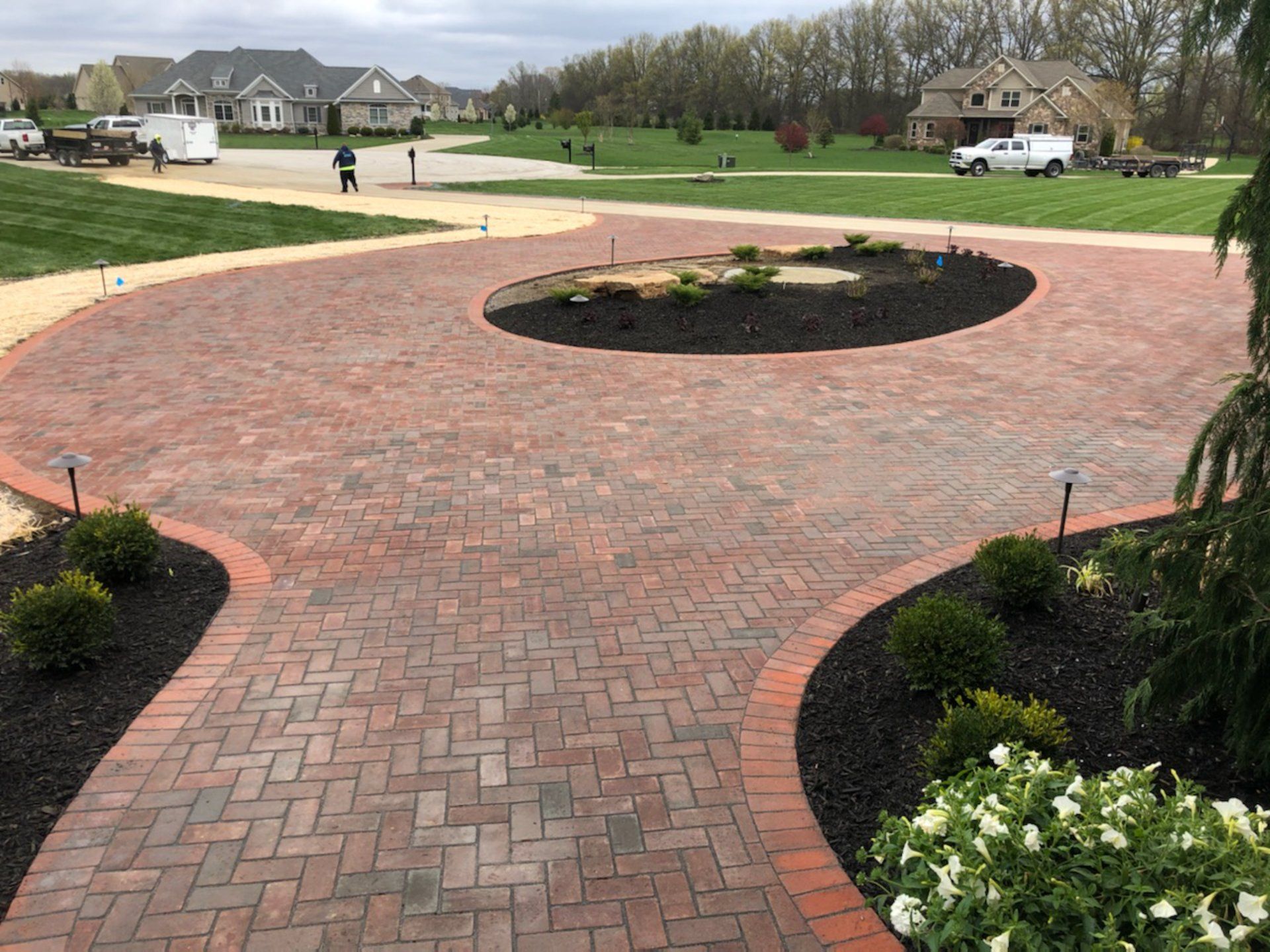 Brick driveway with a circular center, lined with dark mulch, leading to a residential area with houses and landscaping.