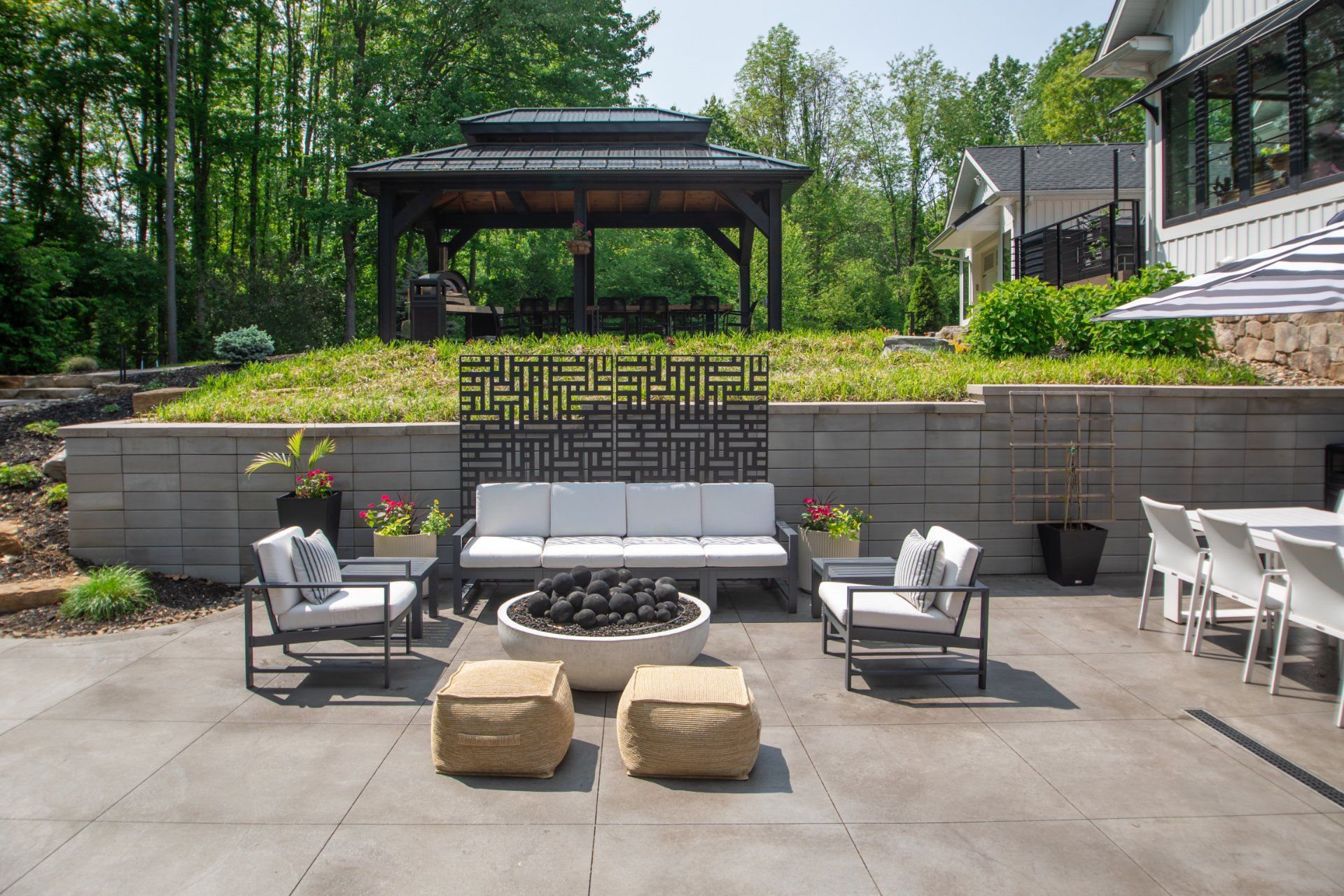 Patio with seating, fire pit, and gazebo. Concrete patio, stone wall with plants, and trees in background.