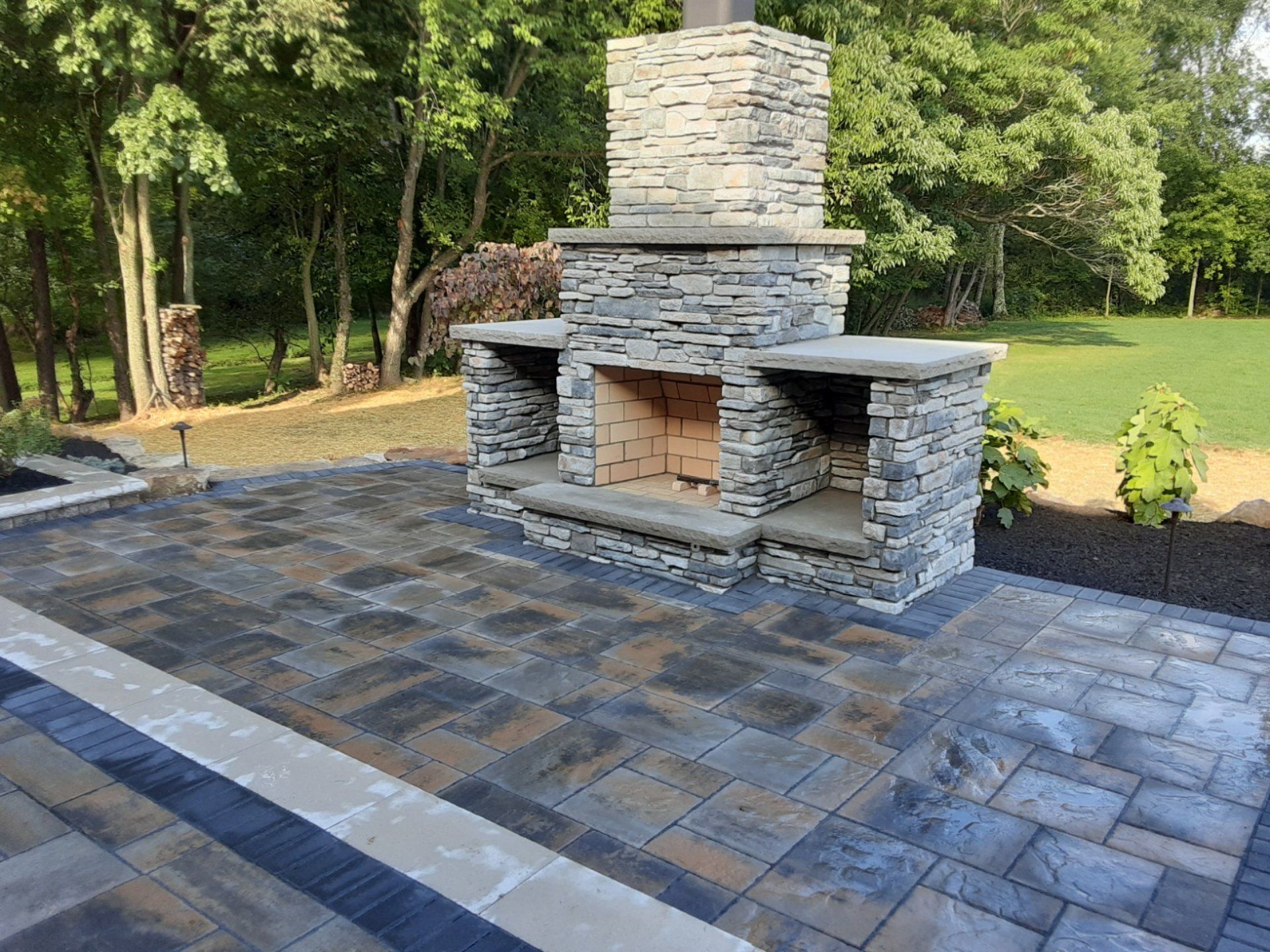 Stone outdoor fireplace with shelves on a paved patio with a grassy background.
