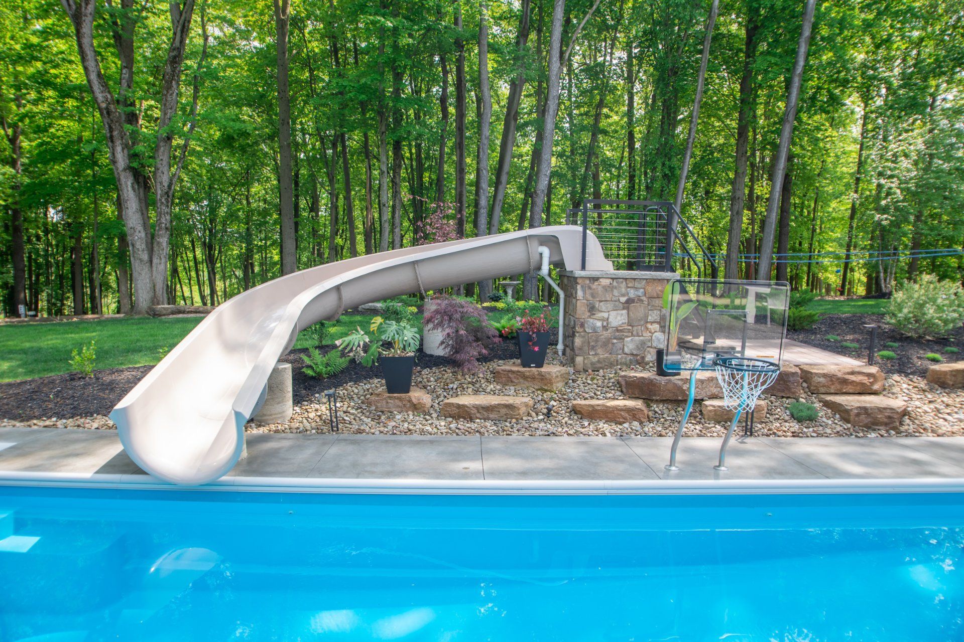 Swimming pool with water slide, stone stairs, and trees in the background. Blue water and light-colored slide.