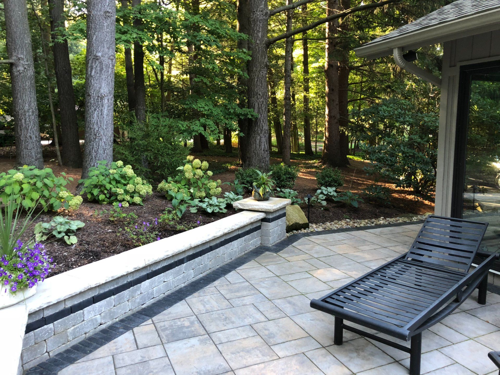 Patio with a lounge chair, landscaping, and a wooded backdrop.