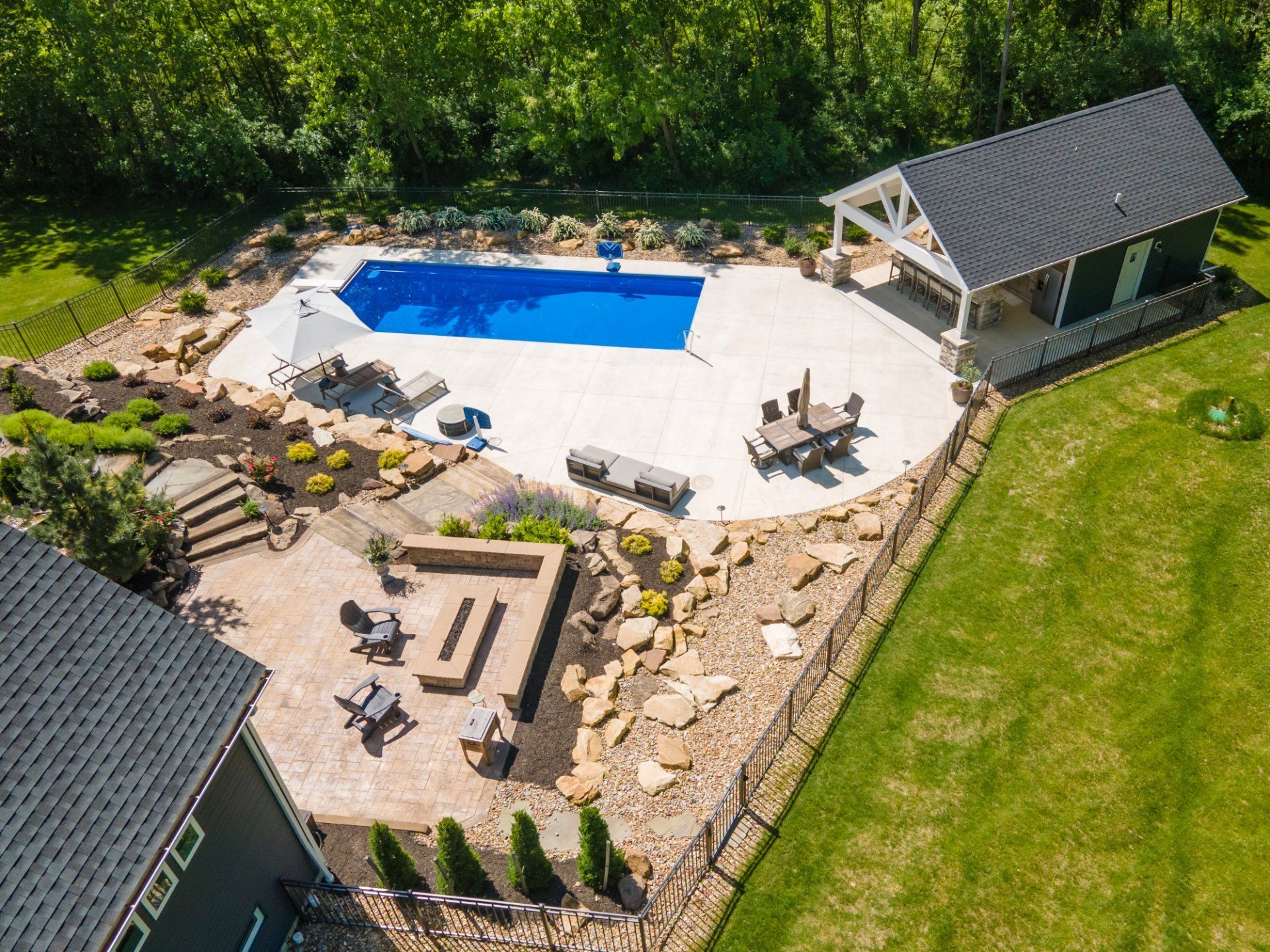 Aerial view of a backyard with a rectangular pool, patio, and a structure with a dark roof. Green grass surrounds.