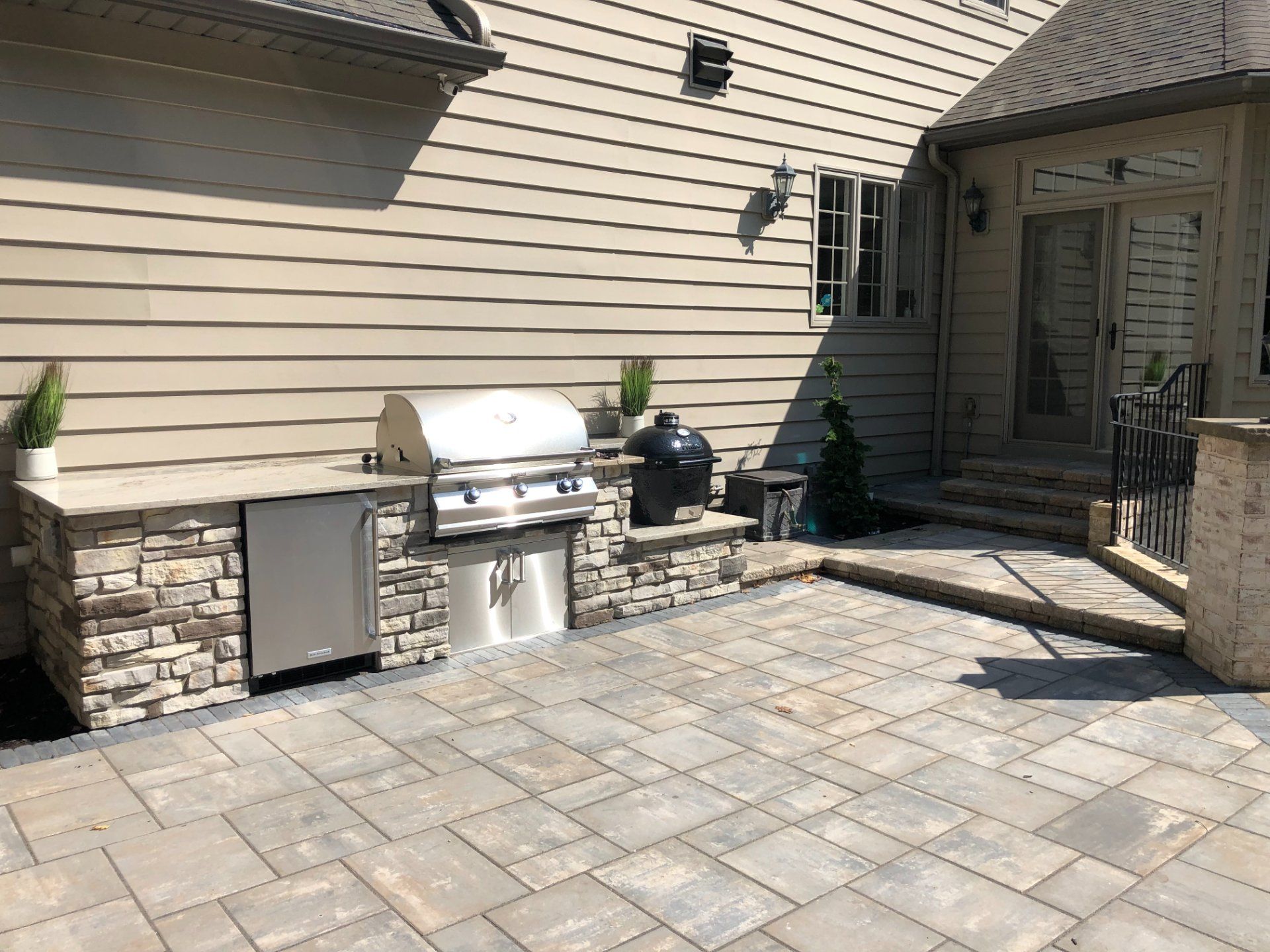 Outdoor kitchen with grill, refrigerator, and smoker on a stone-covered structure. Paver patio connects to a house.