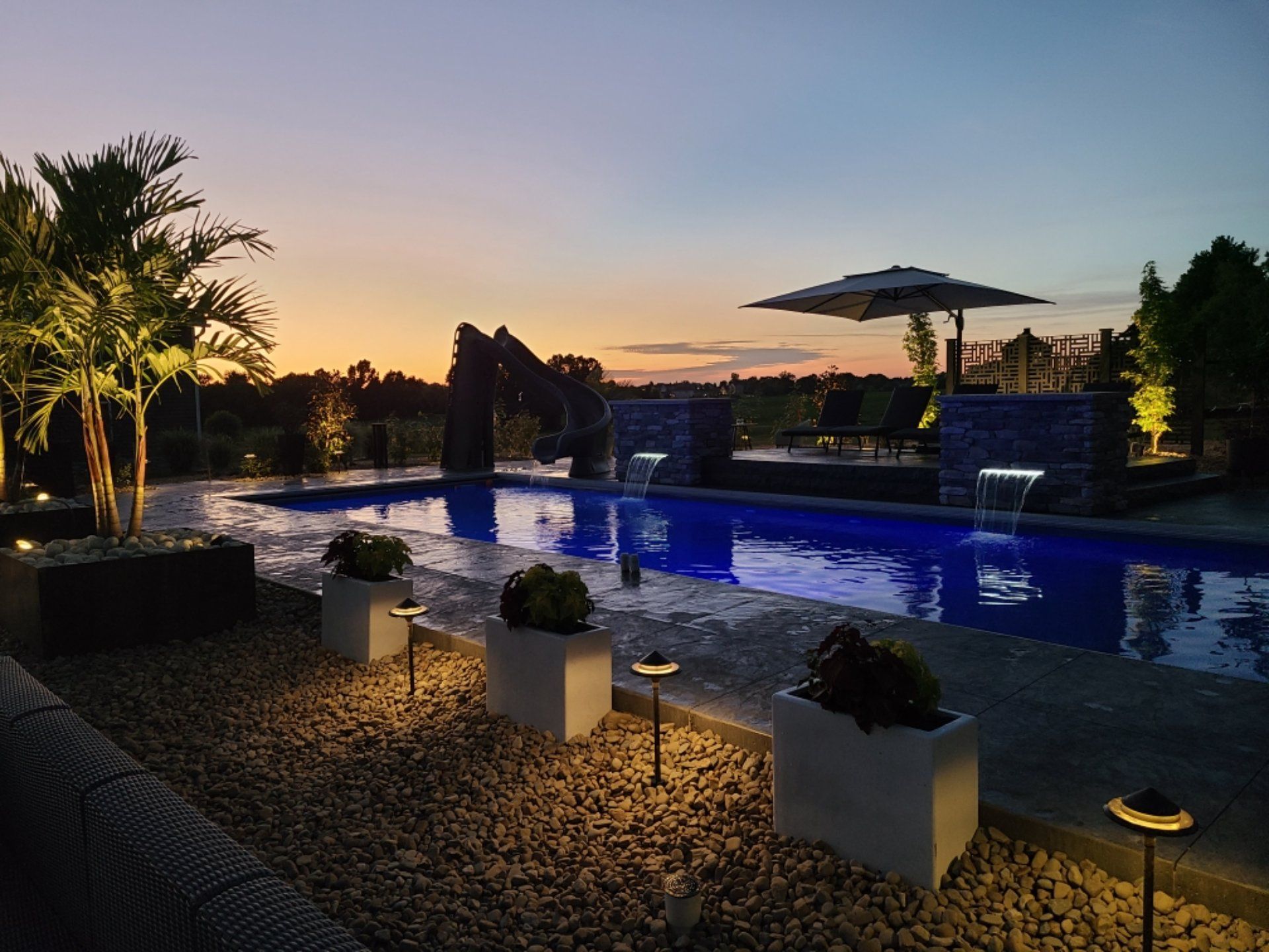 Sunset view of a pool with waterfall features, potted plants, and landscape lighting.