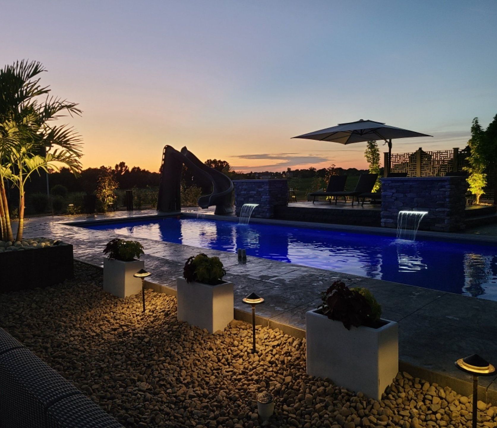 Pool at dusk, illuminated, surrounded by stone and plants. Sunset in the background.