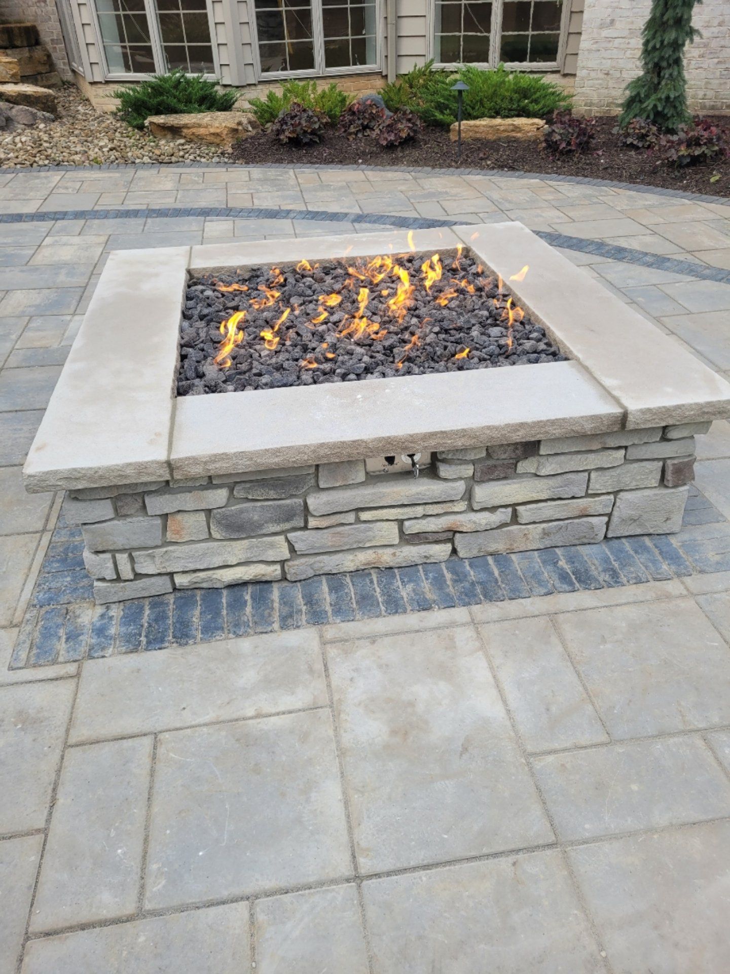 Square stone fire pit with flames, set on a stone patio.