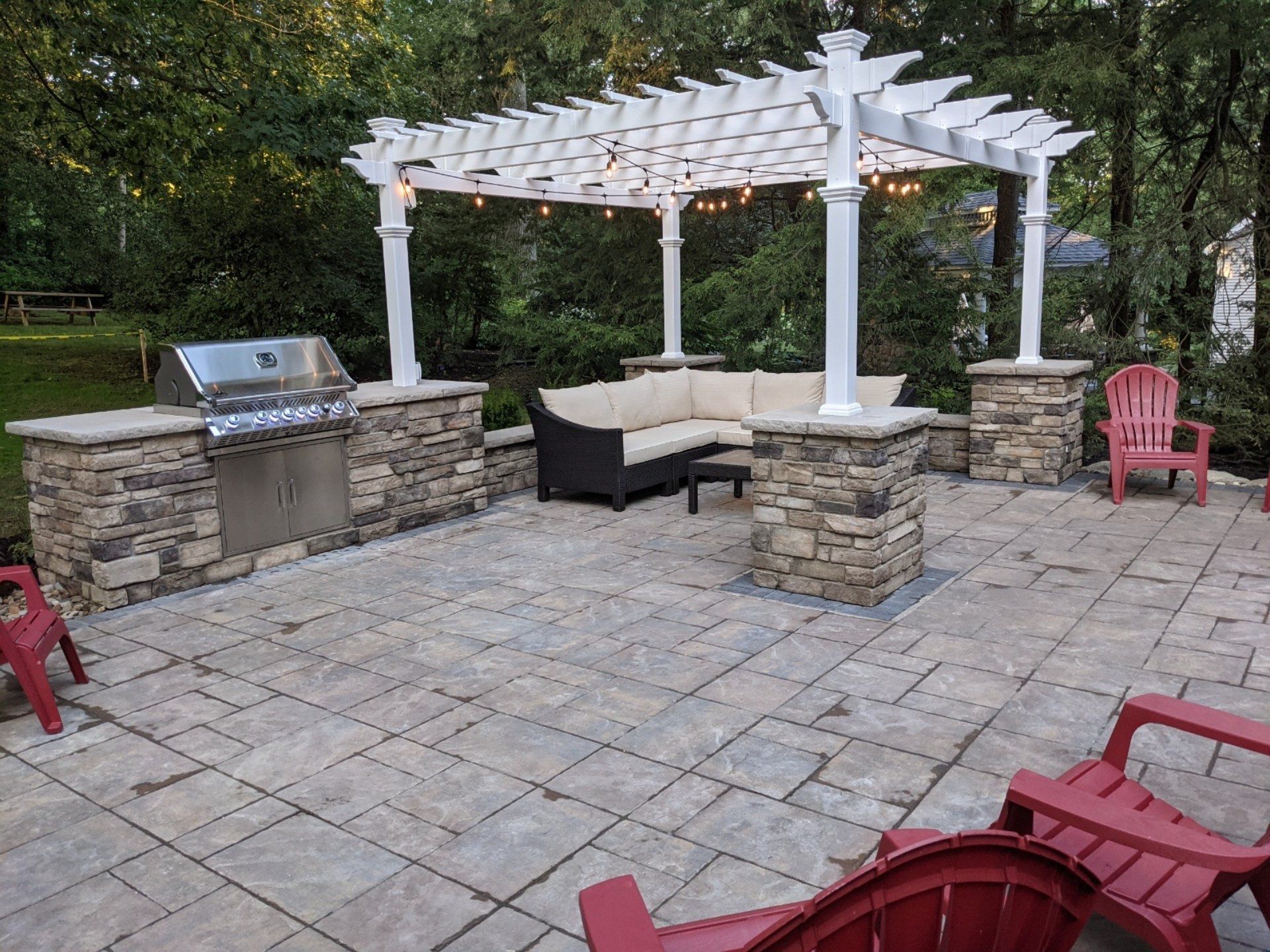 Patio with a grill station, seating, and pergola with string lights, surrounded by trees.