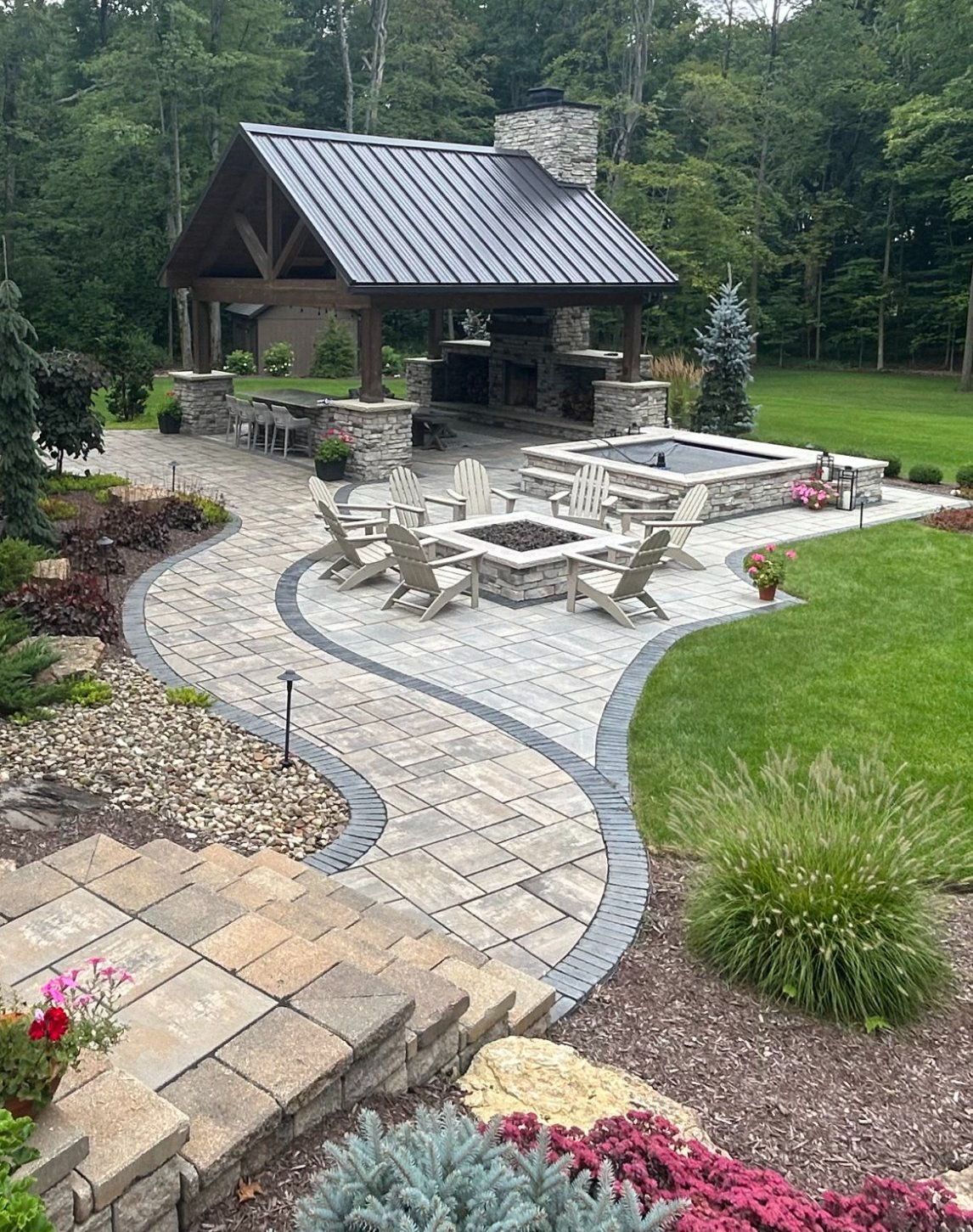 Outdoor patio with stone pavers, fire pit, and covered kitchen area. Green grass and landscaping surround the patio.