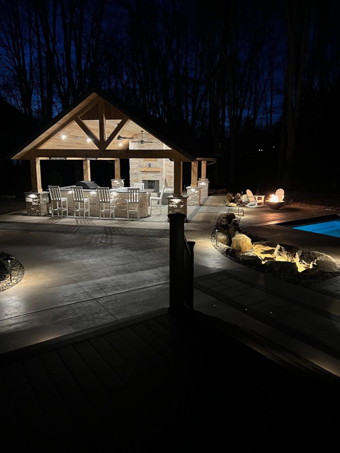 Nighttime view of a patio with a pavilion, grill, seating, and pool. Lit by exterior lighting; a fire pit glows.