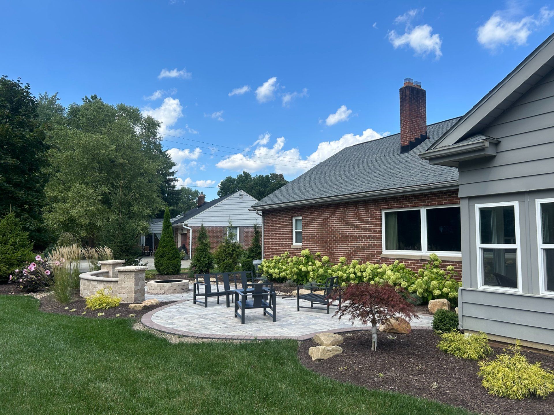 Backyard patio with brick house, gray siding, stone patio, fire pit, seating, green grass, and trees.
