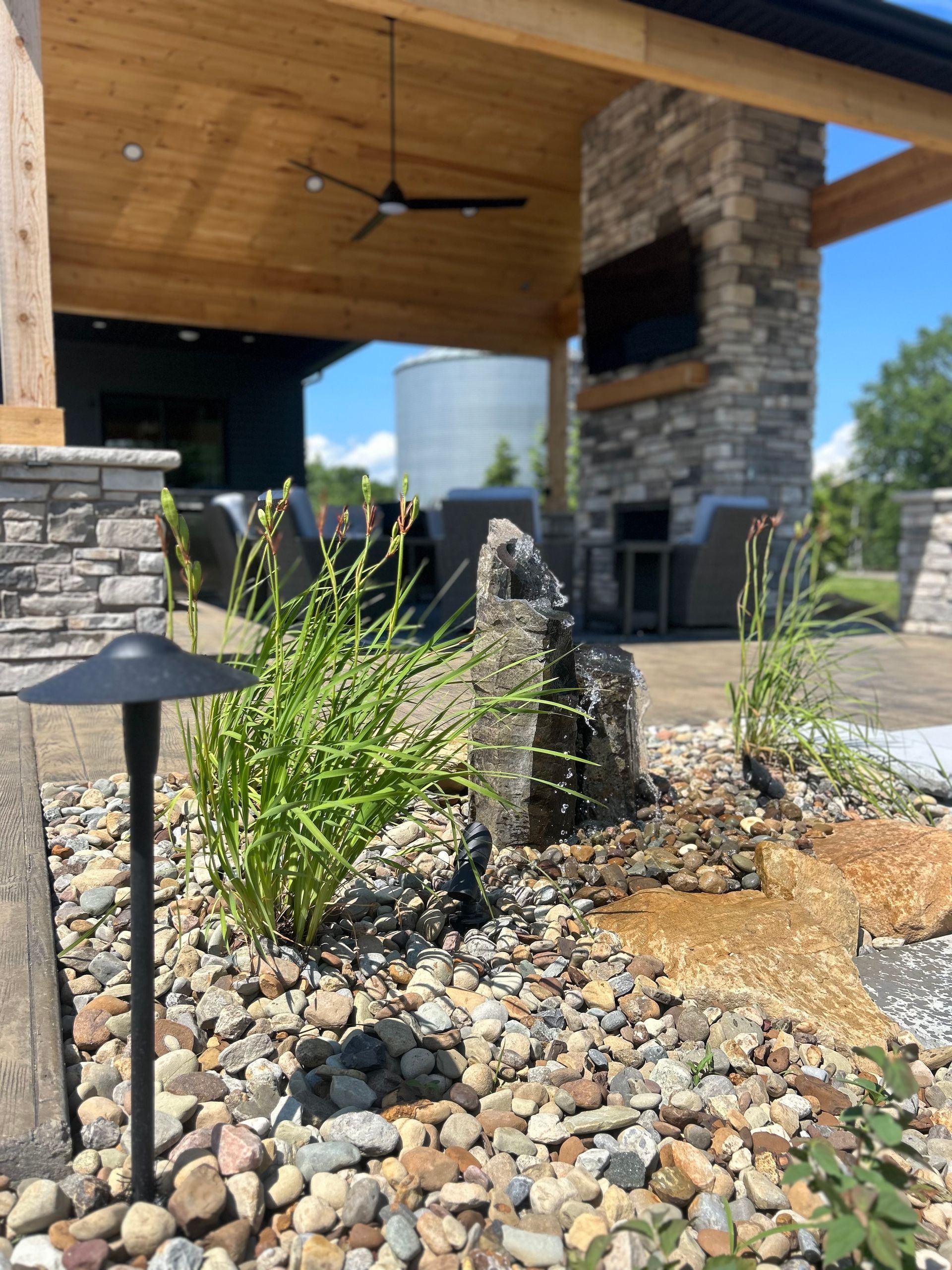 Outdoor patio with rock garden, water feature, and stonework fireplace, under a wood-beamed roof.