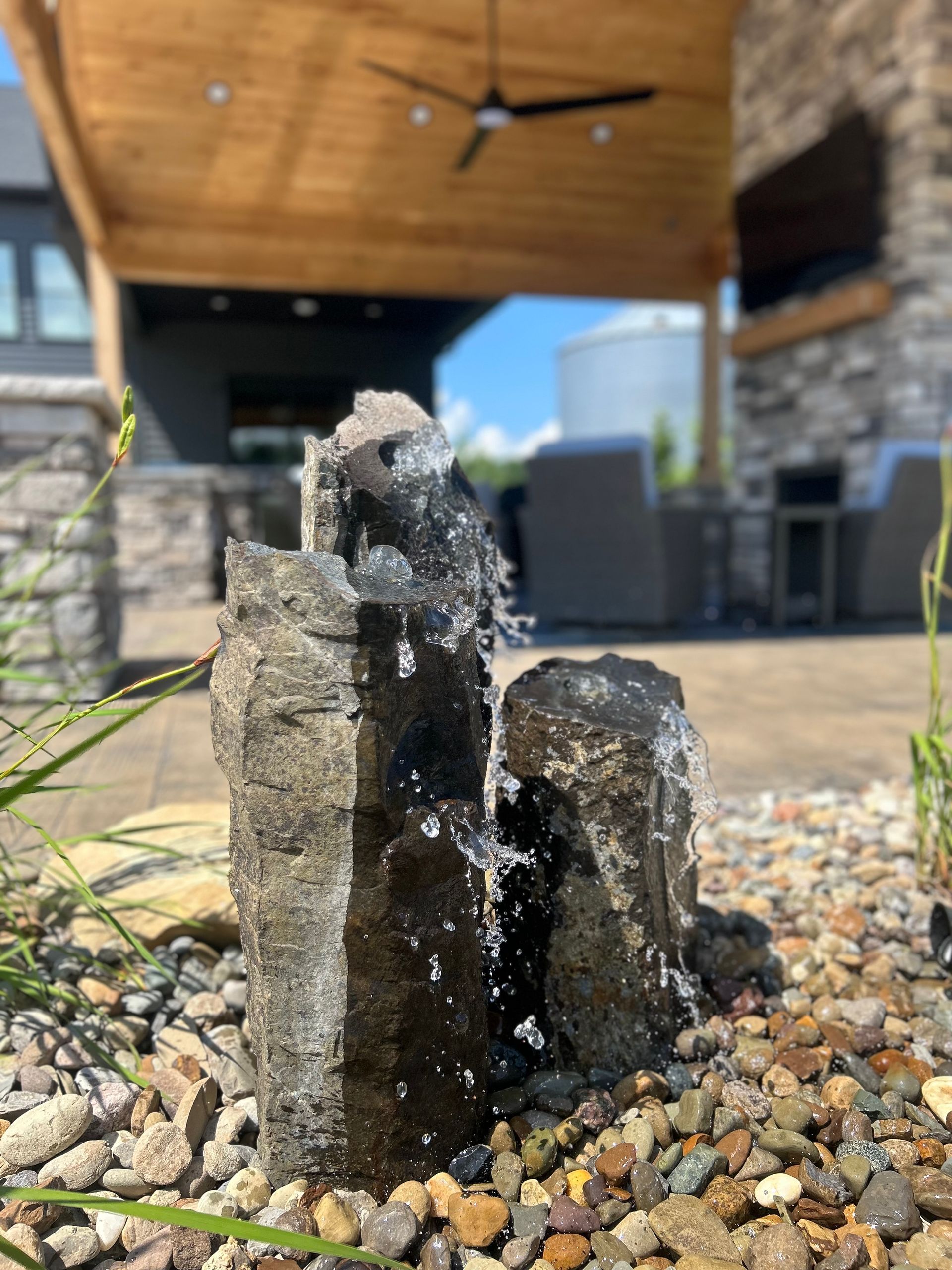 Water fountain made of dark stone, spraying water, set in a stone garden with a blurred patio in the background.