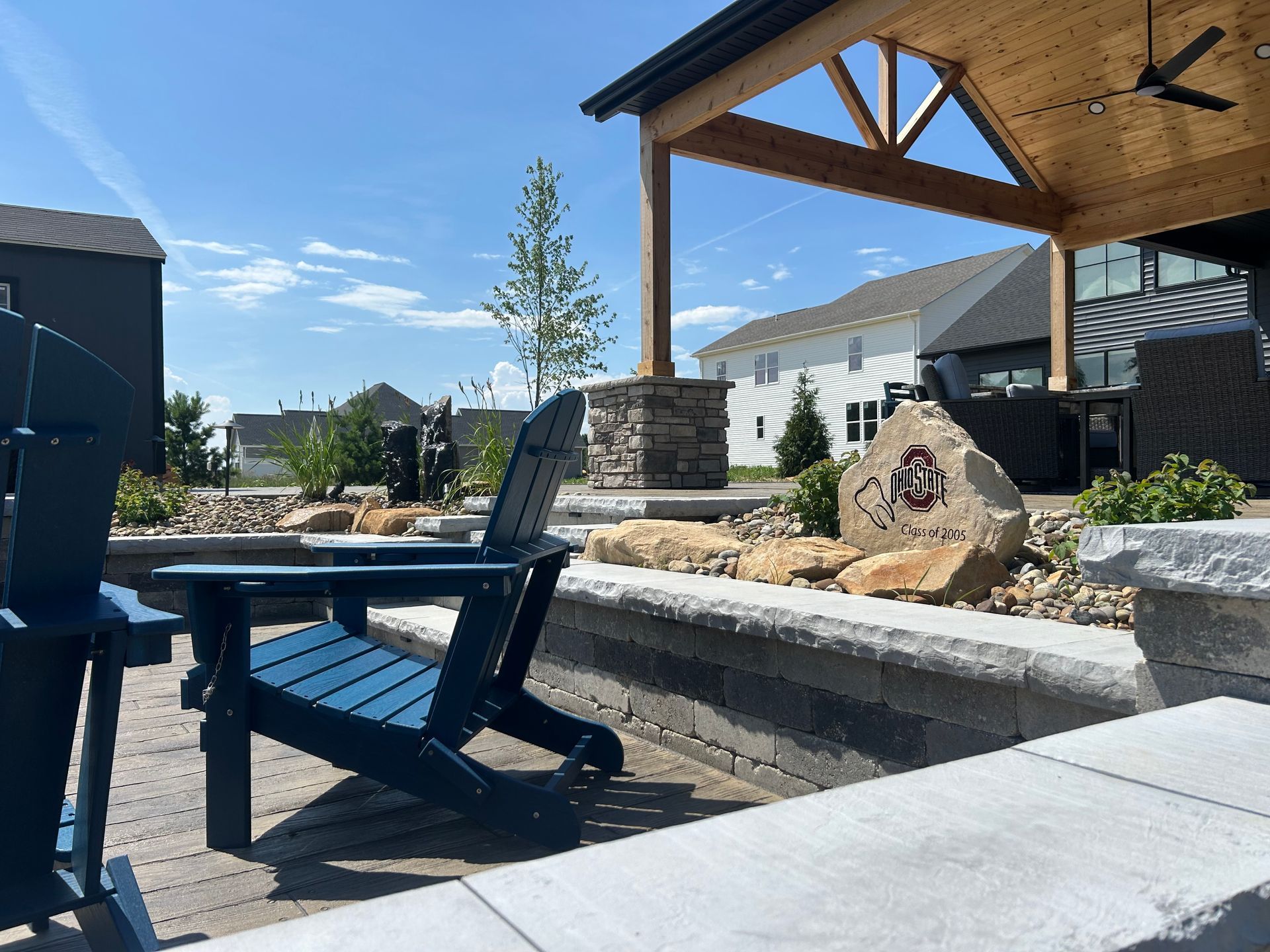 Blue chairs on a patio with a stone fire pit, under a wooden pergola, houses in the background.
