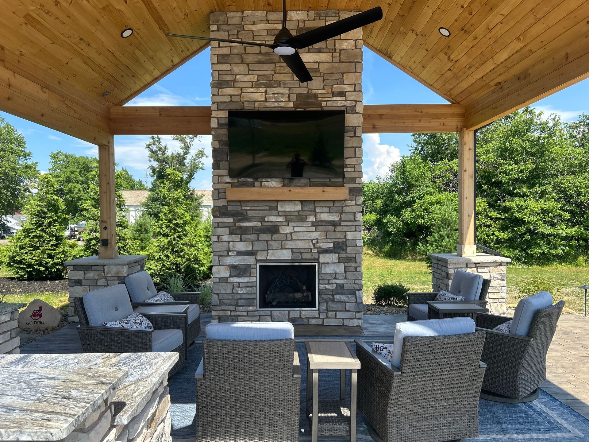 Outdoor patio with fireplace, TV, seating, and overhead fan. Stone and wood construction.