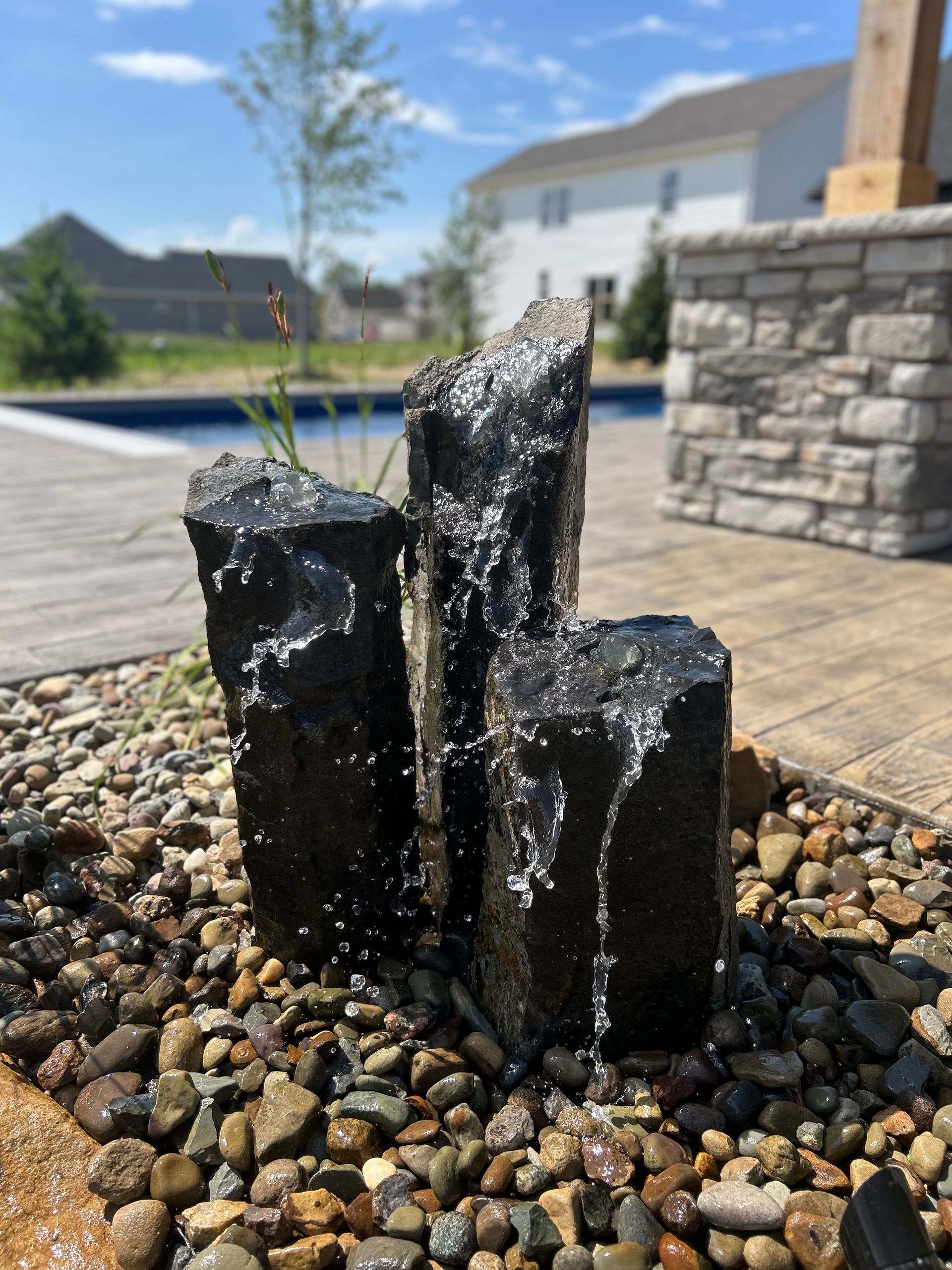 Water fountain with three dark stone columns, water cascading over them, surrounded by pebbles.