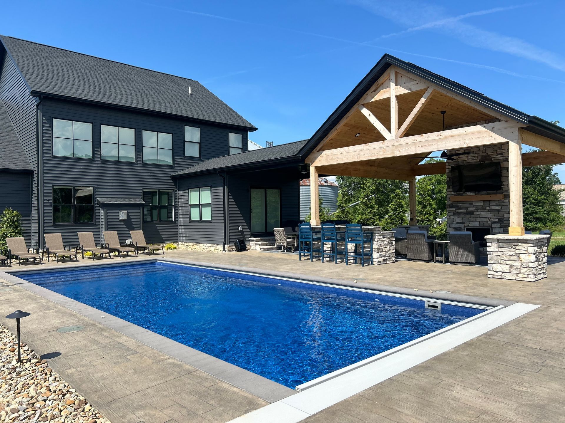 Backyard with a dark house, pool, and covered patio with fireplace. Sunny day, blue water.